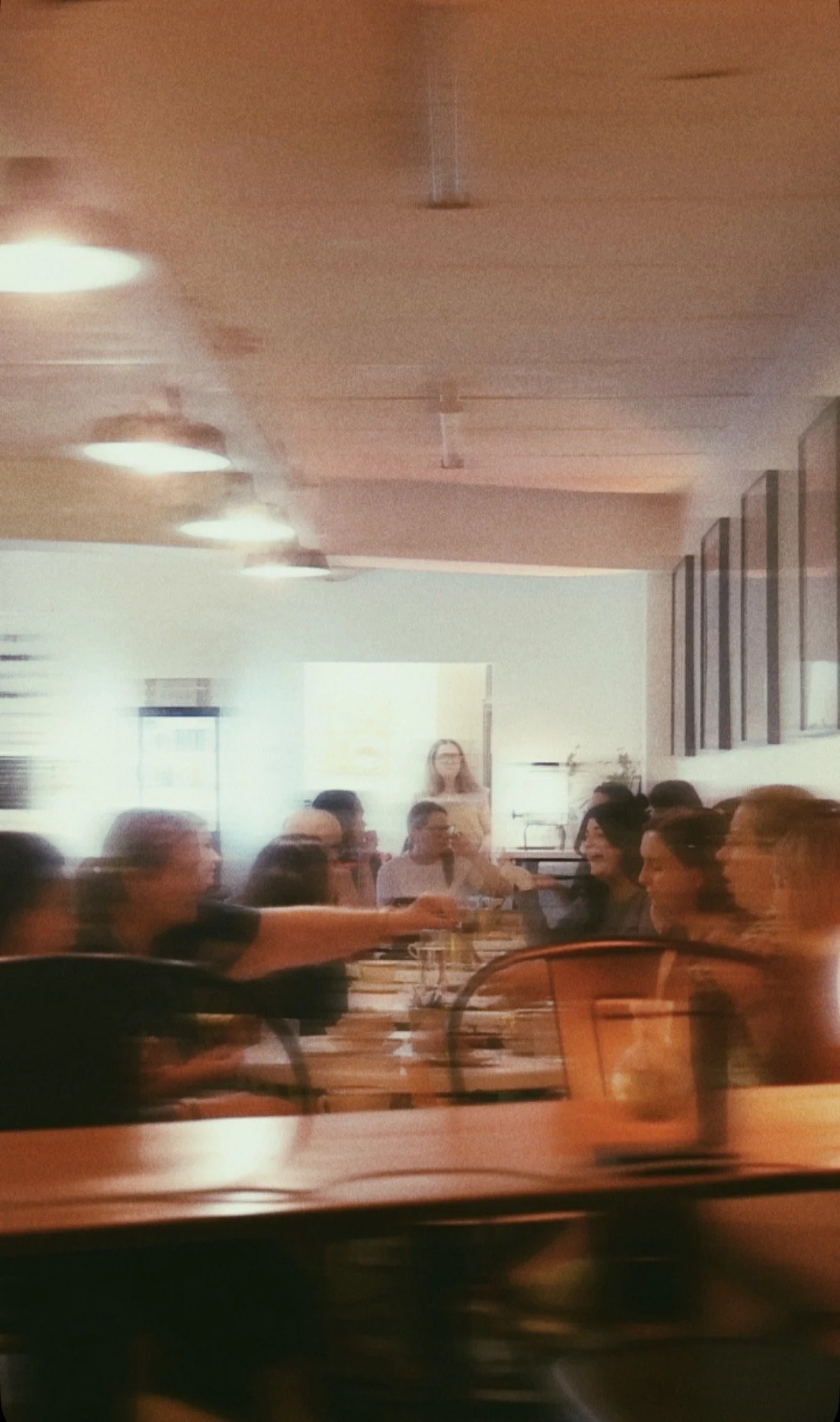 A group of people sitting around a table in a restaurant, with a woman standing in the background, visible through a blurry and warm-colored photo.