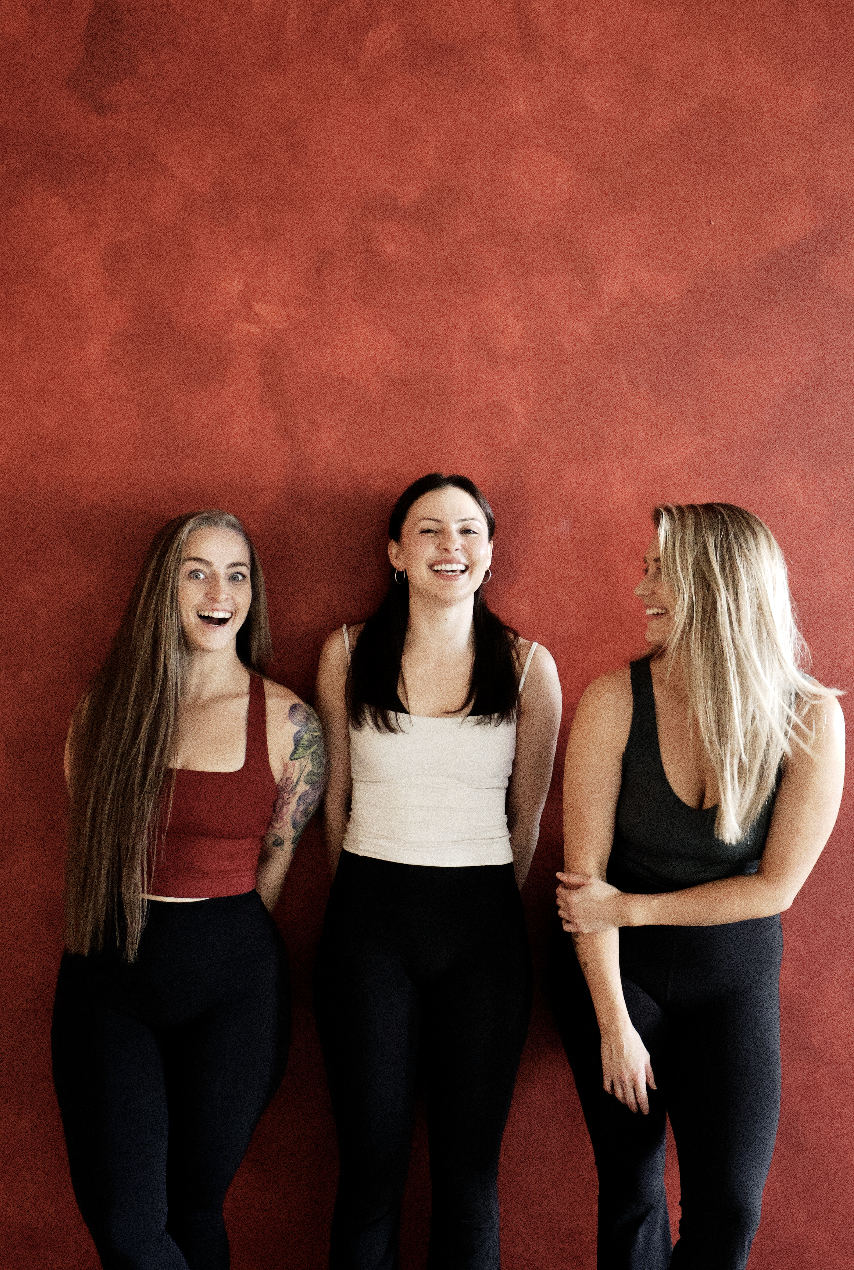 Three women standing against a red wall, smiling and laughing, dressed in casual tops and black pants.