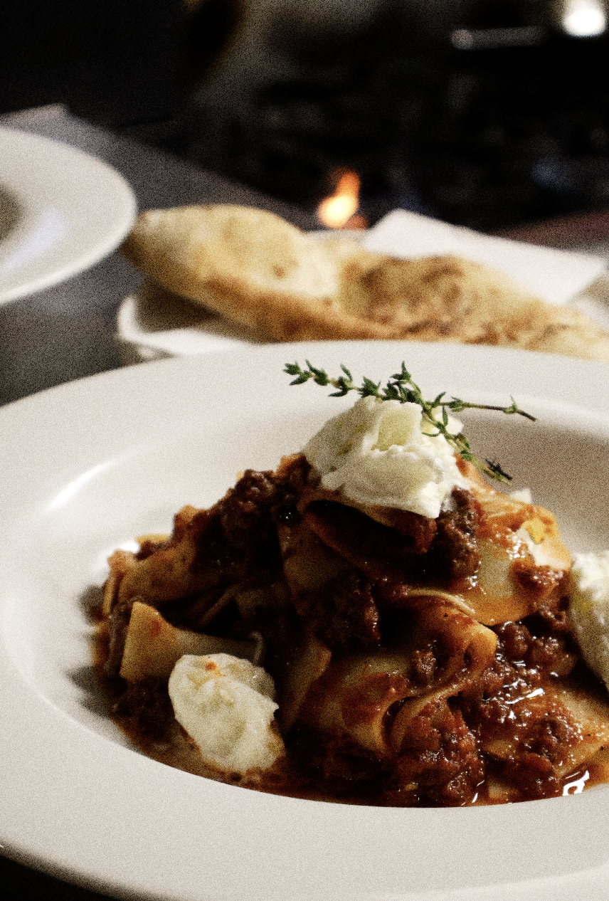 A plate of lasagna topped with a dollop of ricotta cheese and a sprig of thyme, with a piece of naan bread in the background.