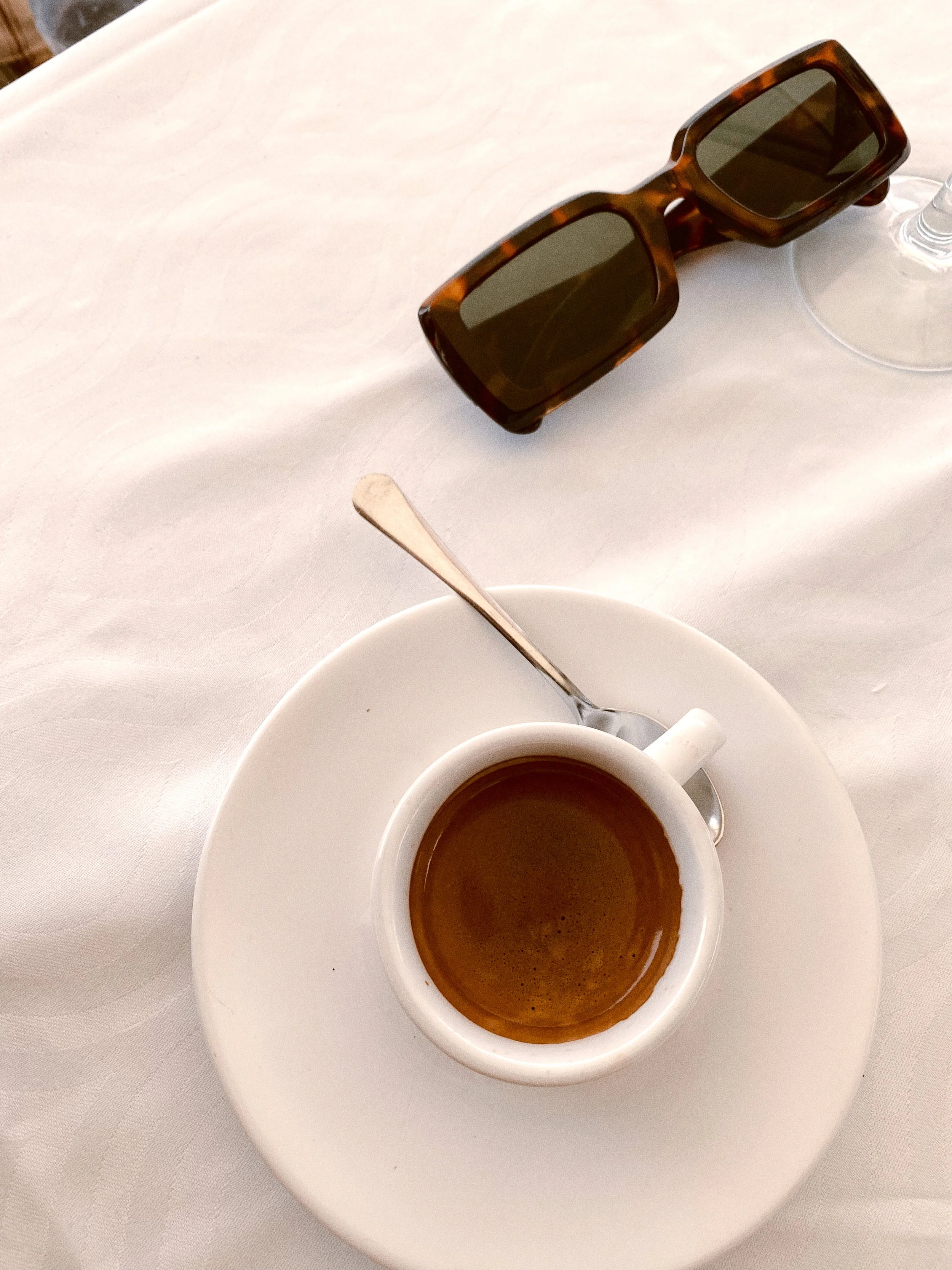A white cup of hot coffee on a saucer with a silver spoon, on a white tablecloth, with a pair of sunglasses nearby.