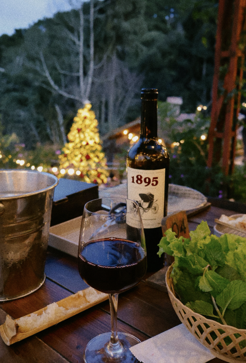 A glass of red wine on a wooden table outdoors at dusk with a bottle labeled 1895, a basket of green lettuce, a bucket, and a small wooden sign with the number 14. In the background, there is a blurred Christmas tree decorated with lights and red orn