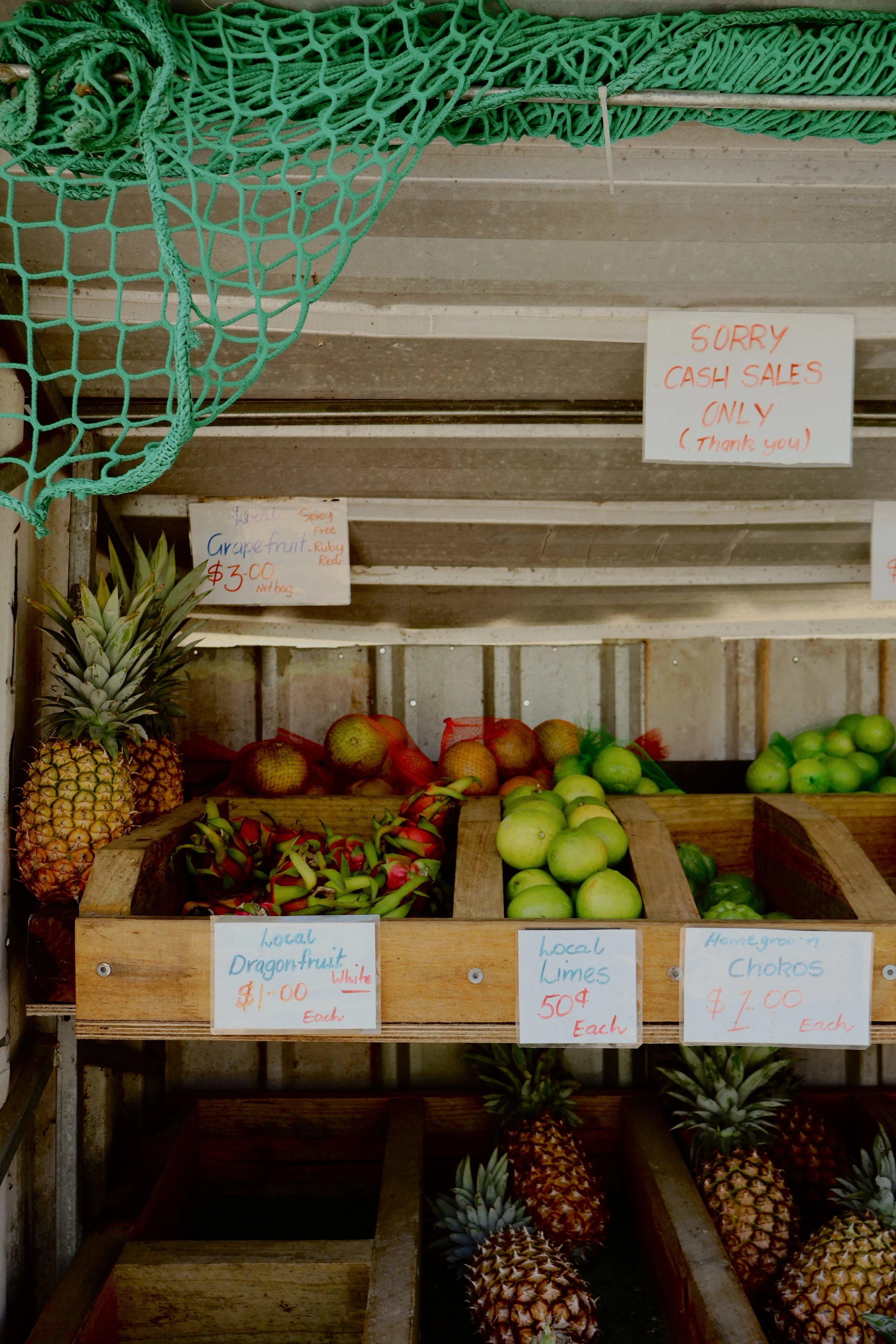 Display of fresh pineapples, apples, dragon fruit, and limes in wooden baskets at a market stand with signs indicating prices and product information.
