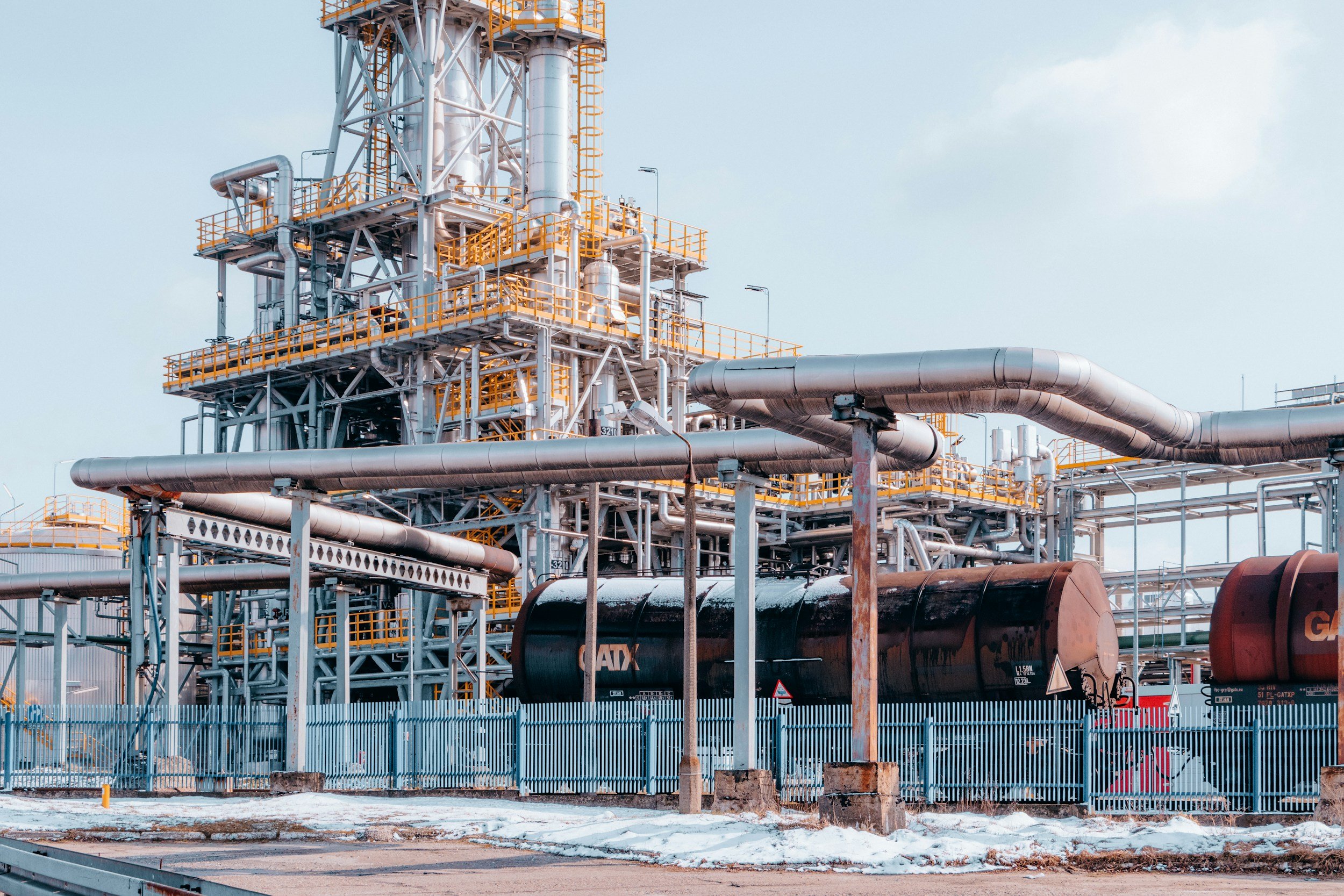 Industrial refinery with metal pipes, tanks, and yellow safety railings, surrounded by a blue fence, with snow on the ground. Source: Unsplash