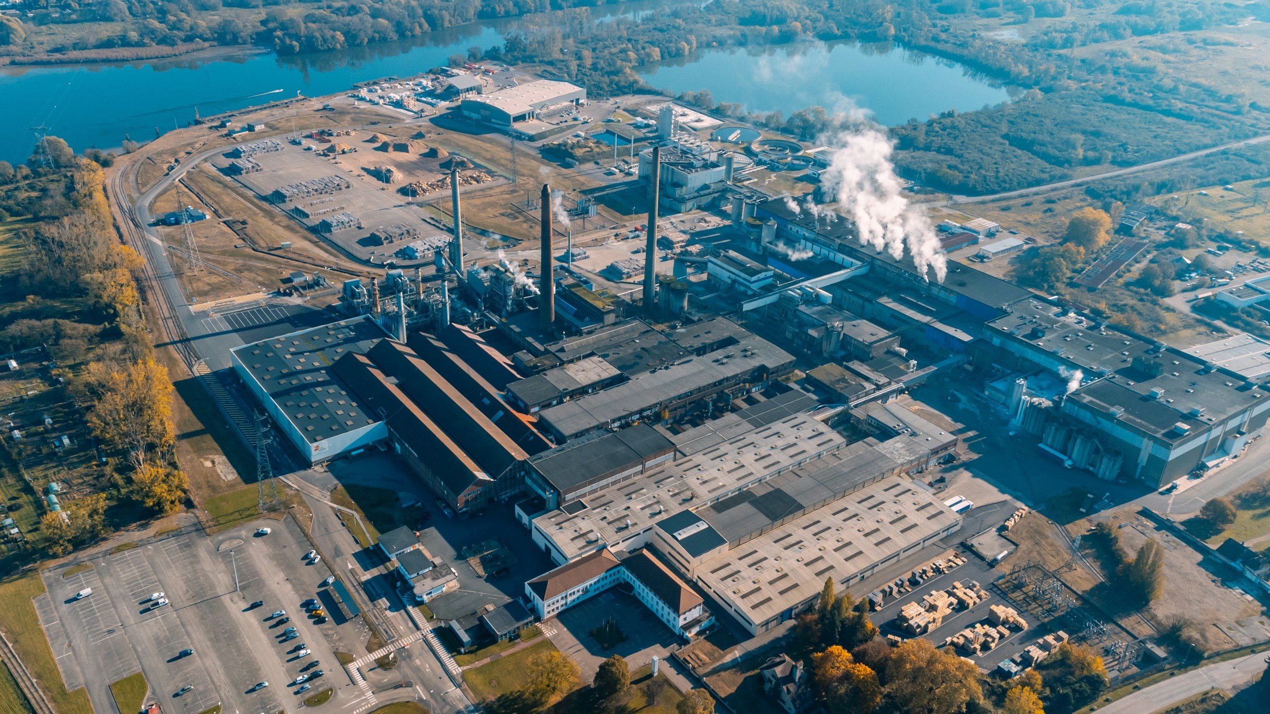 Aerial view of an industrial factory complex with smokestacks emitting steam, surrounded by parking lots, roads, and greenery, near a body of water. Source: Unsplash