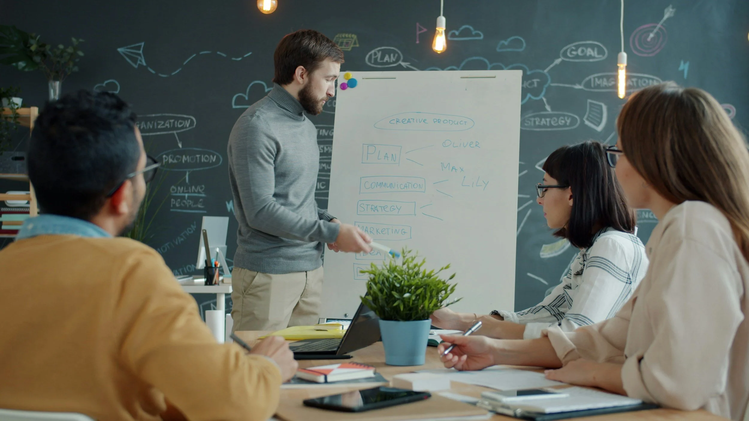 Business meeting with four people in a conference room, a man presenting ideas on a whiteboard, and three women listening and taking notes.  Source:Unsplash