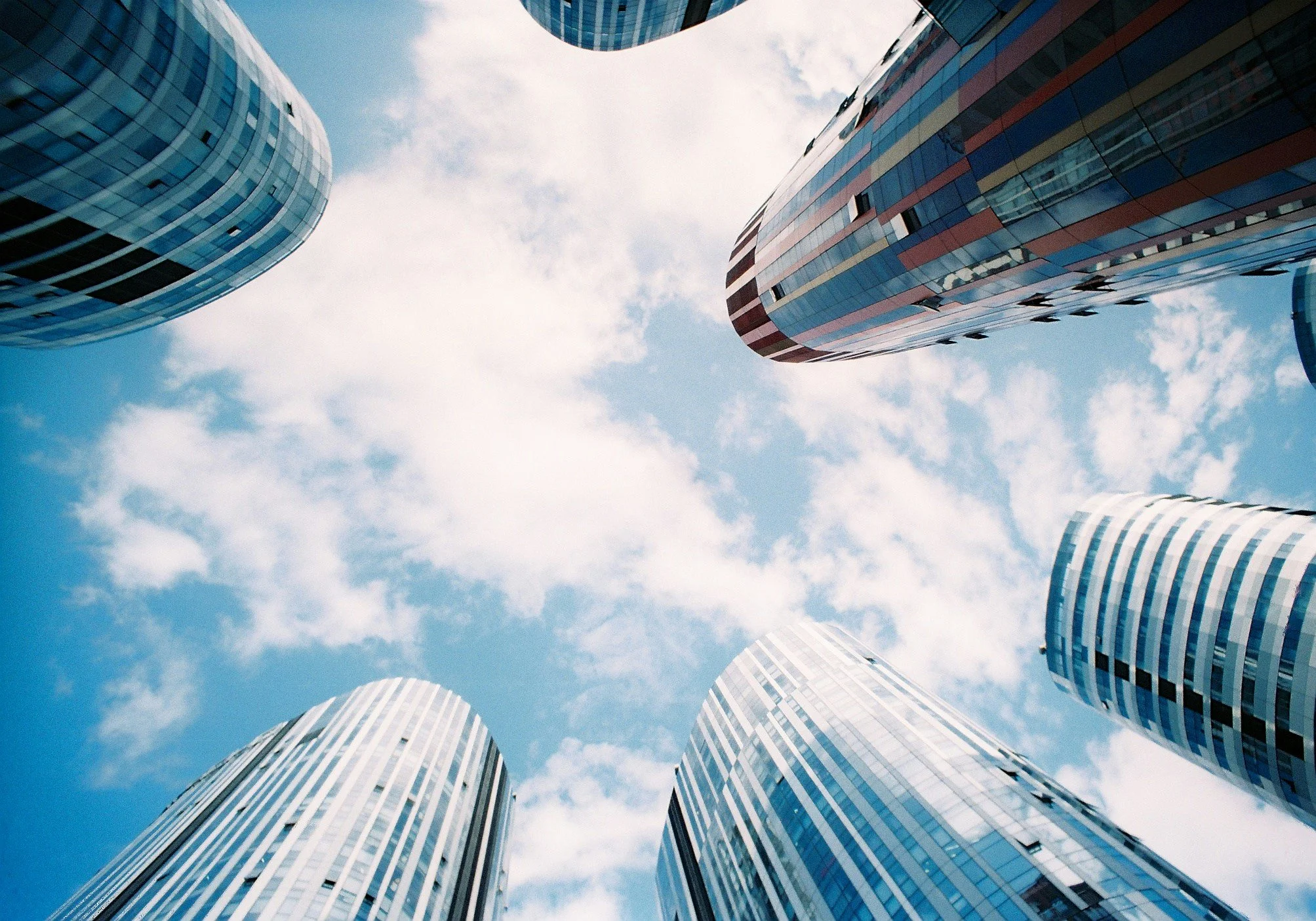 Looking up at modern high-rise office buildings with glass facades against a blue sky with scattered clouds.