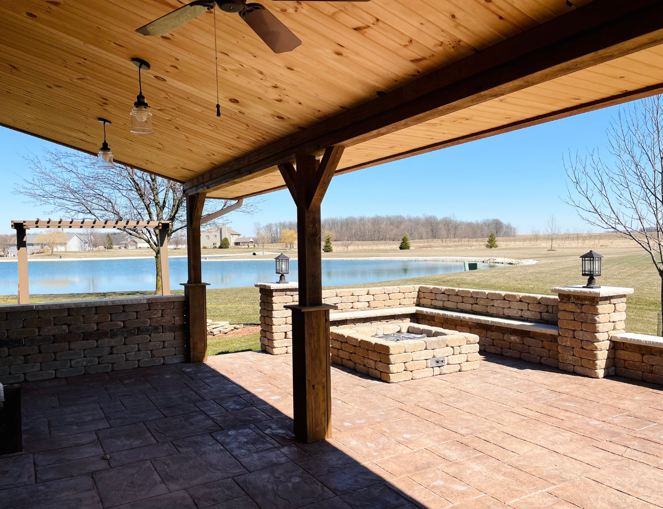 Covered patio overlooking a pond with a stone fire pit and brick benches, surrounded by open grassy fields and a few trees, under a clear blue sky.