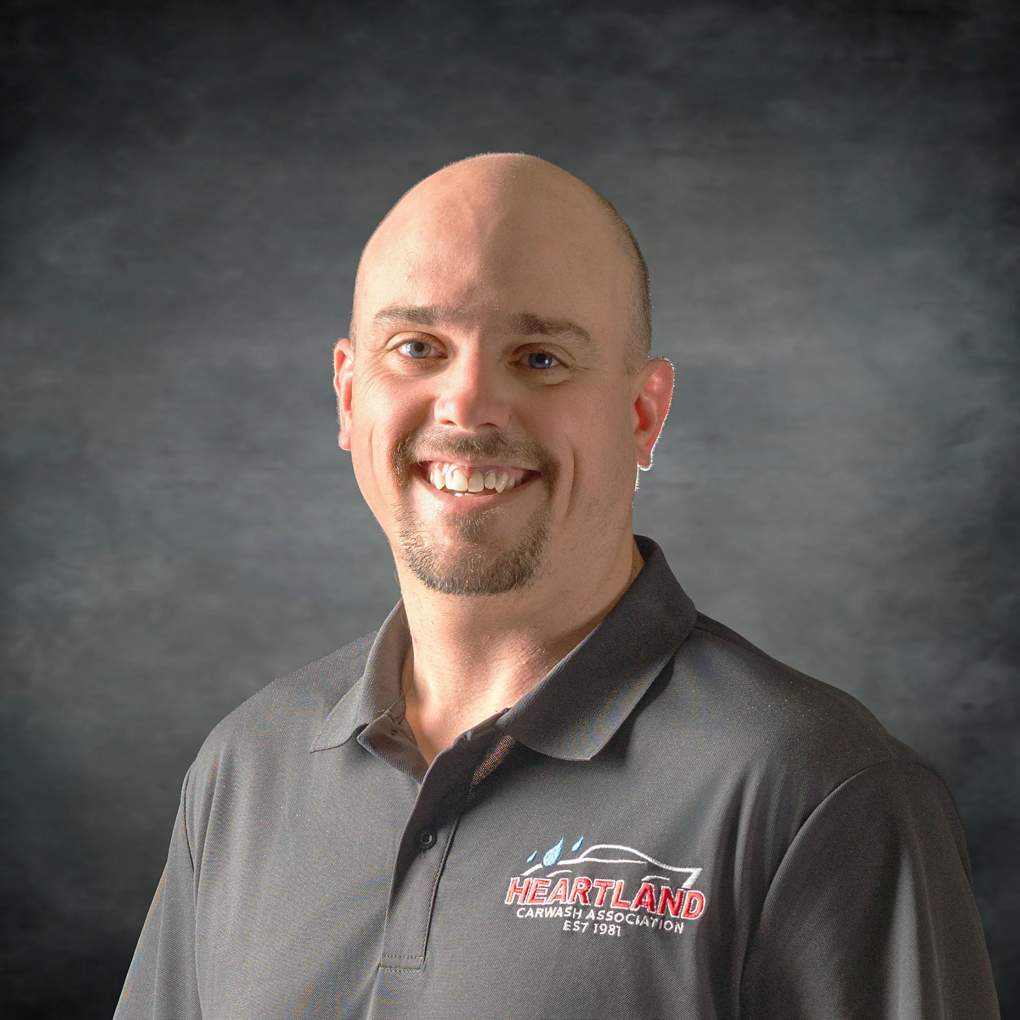 Close-up of a smiling man with a shaved head, wearing a black shirt with a Hartland Heartland Carvings Association logo, against a dark background.