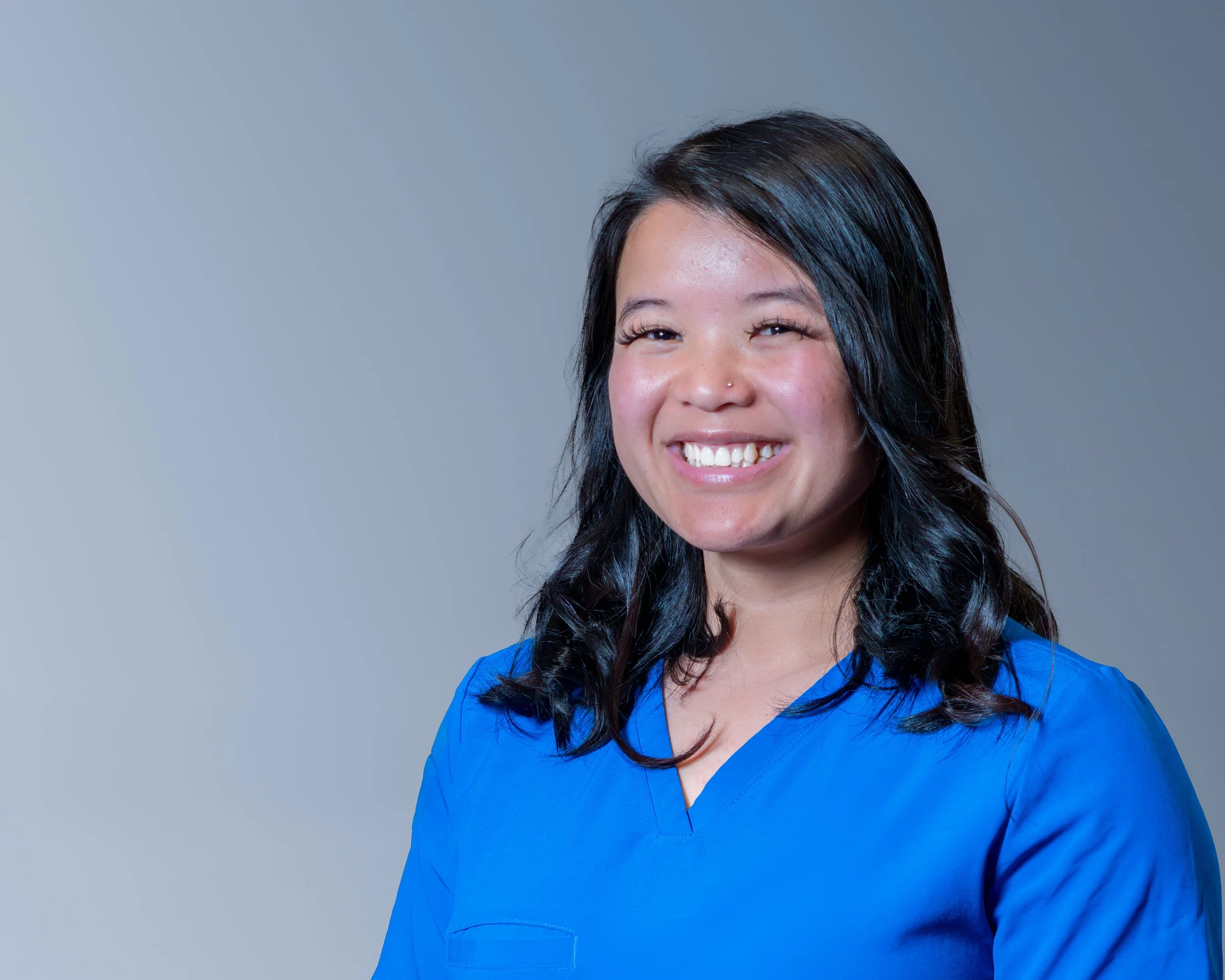 Close-up of a smiling woman in blue scrub top against a plain gray background.