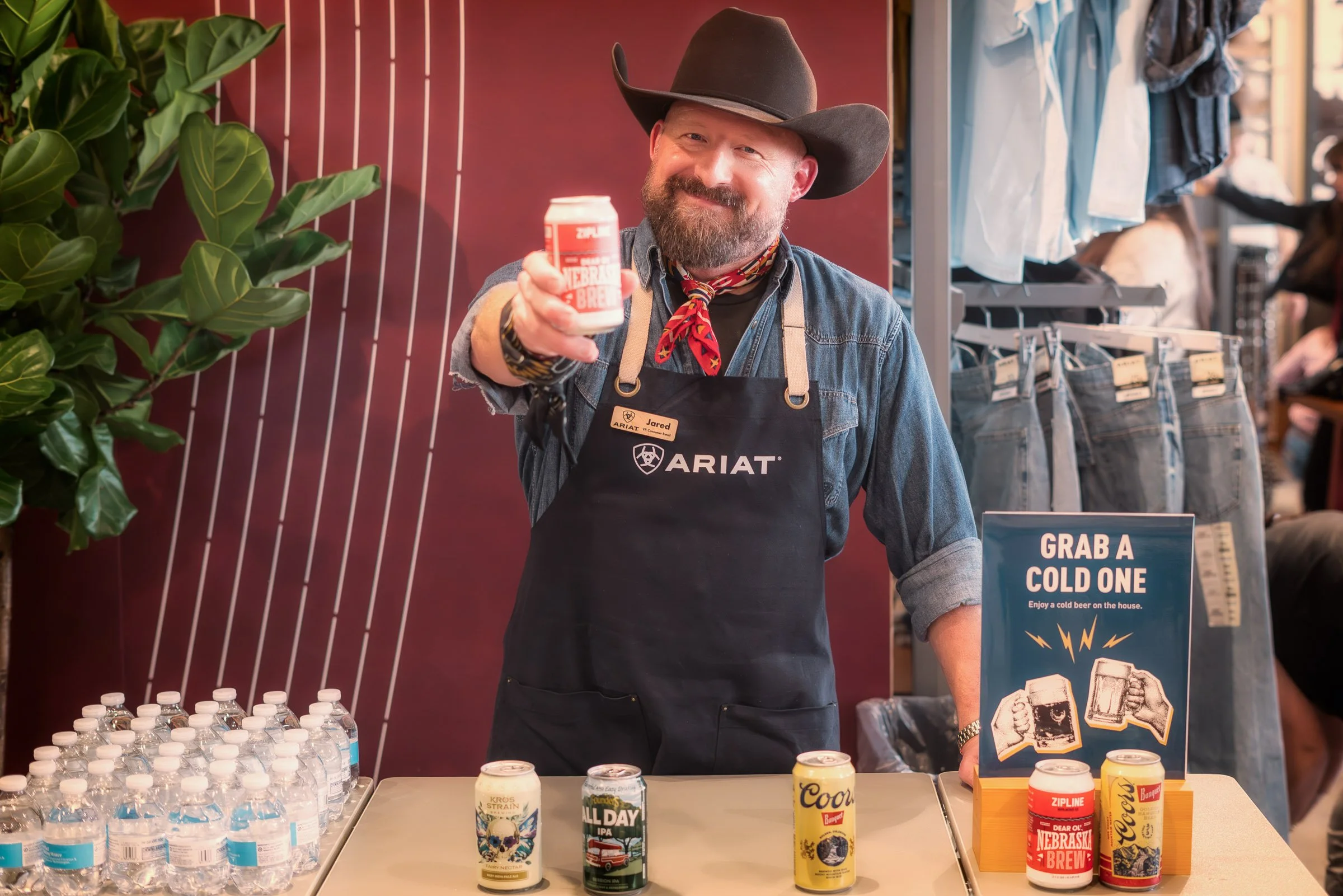 Man wearing a cowboy hat and an apron smiling and holding a can of Nebraska brew beer inside a store or brewery. There are bottled water, canned drinks, and promotional signs on the table in front of him. Shelving with clothes is visible in the backg
