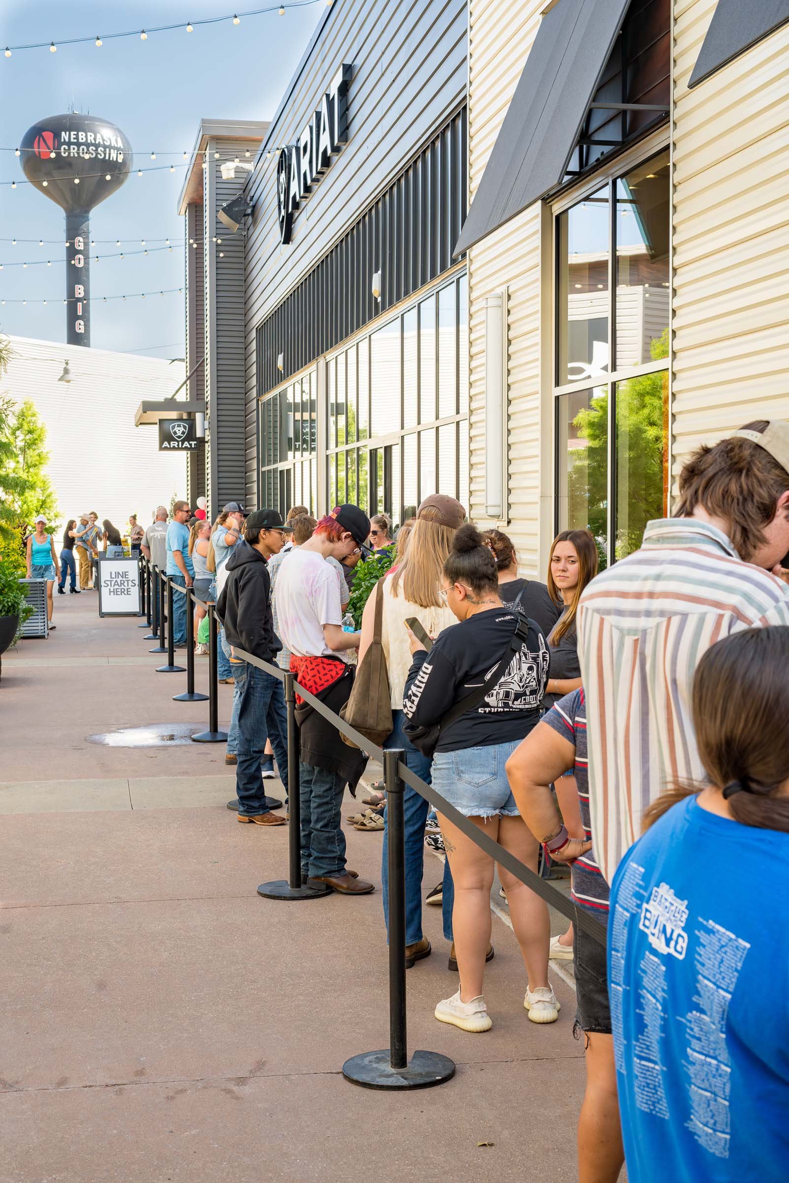 People waiting in line outside a building with large windows, with a sign indicating the line starts there, near a water tower with Nebraska Crossings written on it.