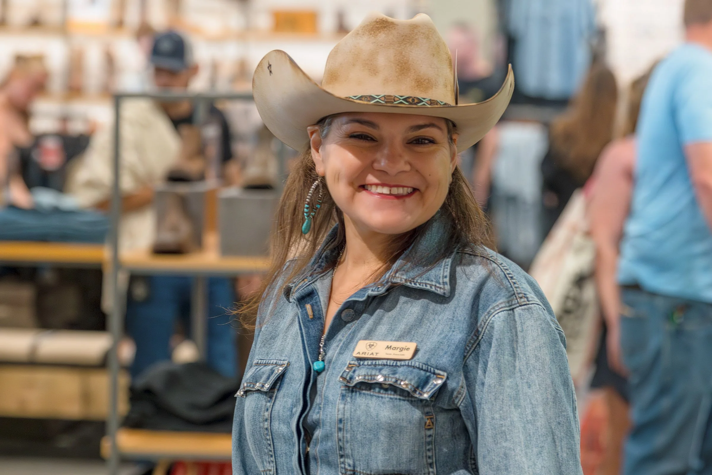 A woman smiling wearing a cowboy hat and denim jacket with a name tag that says Margie at an indoor event with blurred crowd and shelves in the background.