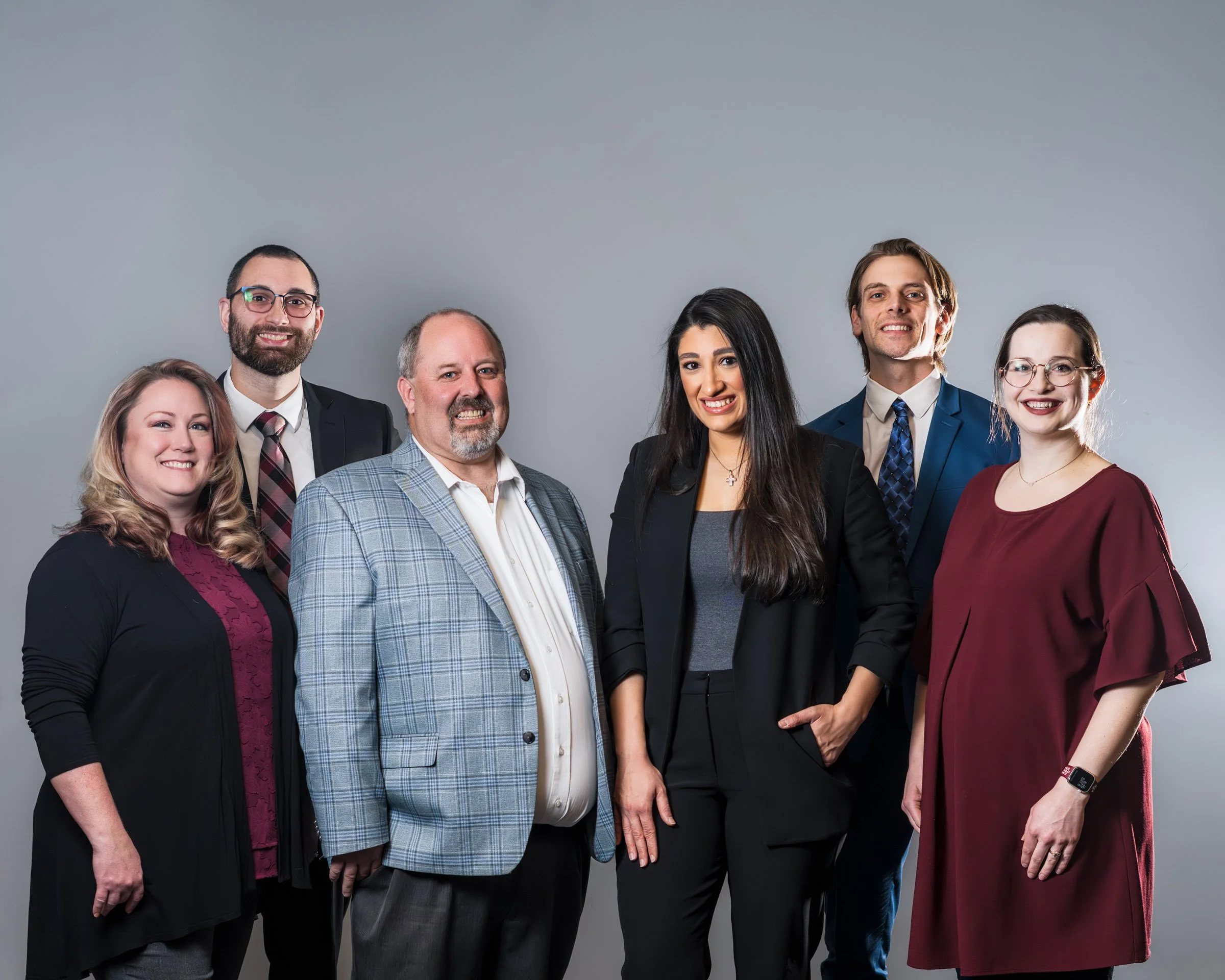 Six diverse professionals standing together against a gray background, smiling, dressed in business attire.