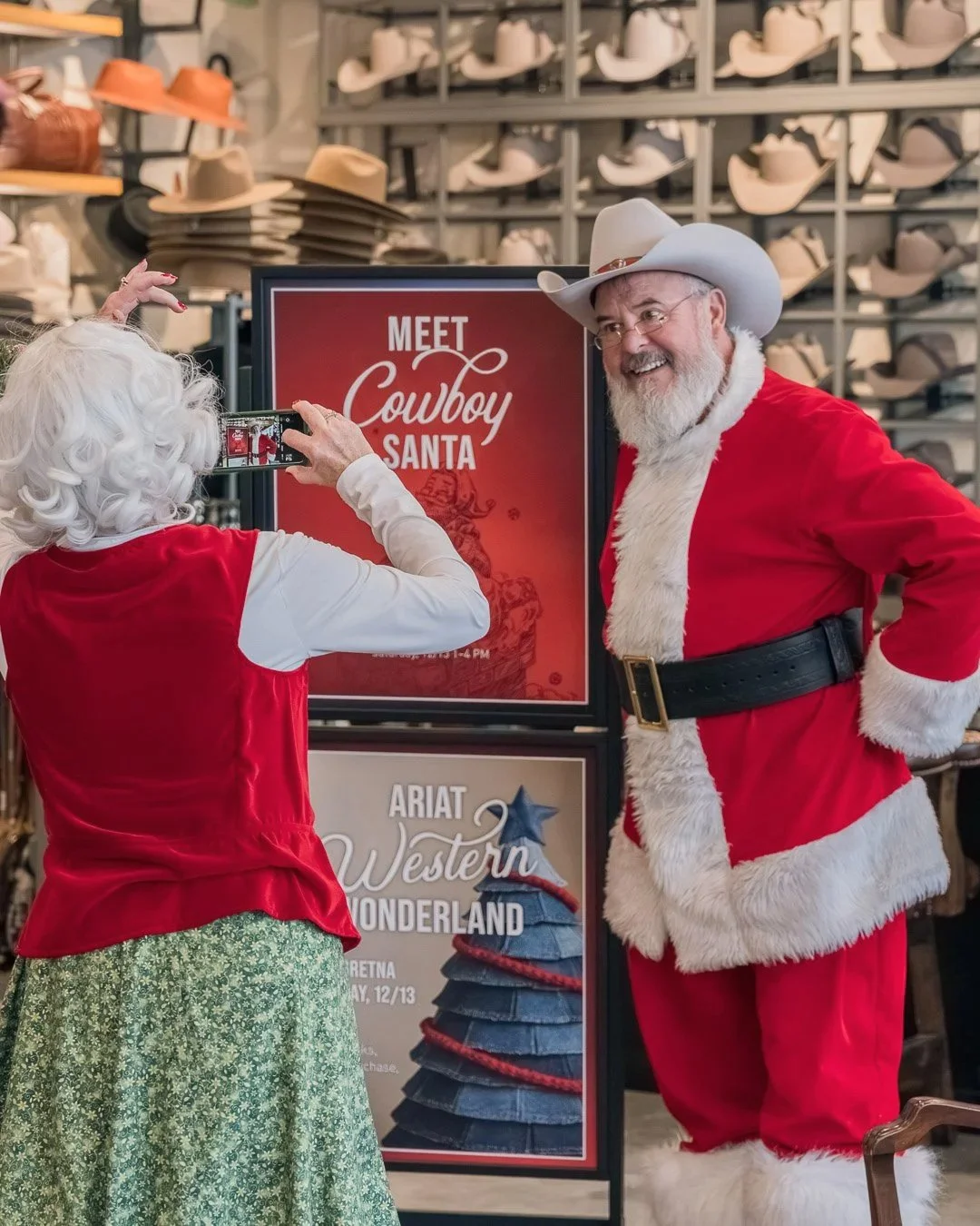 A man dressed as Santa Claus and a woman, seen from the back, taking a photo of Santa with a woman dressed as a cowgirl with white curly hair. They are in a store with shelves of hats in the background.