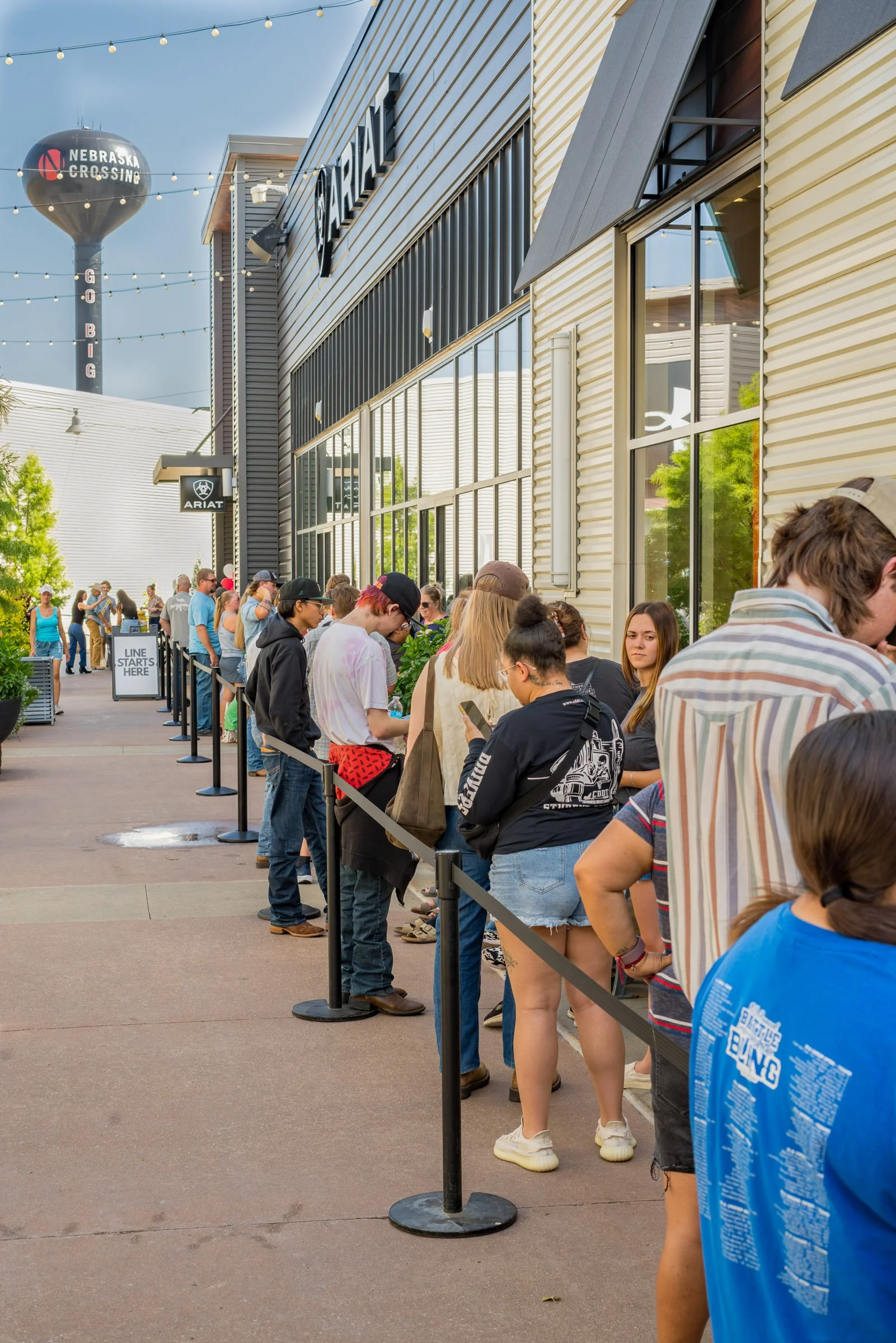 People lined up along a sidewalk outside a building, waiting in a single file behind a queue barrier with a sign that says 'Line Starts Here'. The building has large windows and modern architecture. There are trees and a cloudy sky, with a water towe