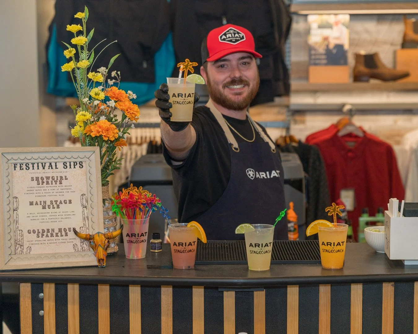 Smiling bartender wearing a black apron with ARIAT logo, red cap, and gloves, serving colorful cocktails with garnishes at a bar.