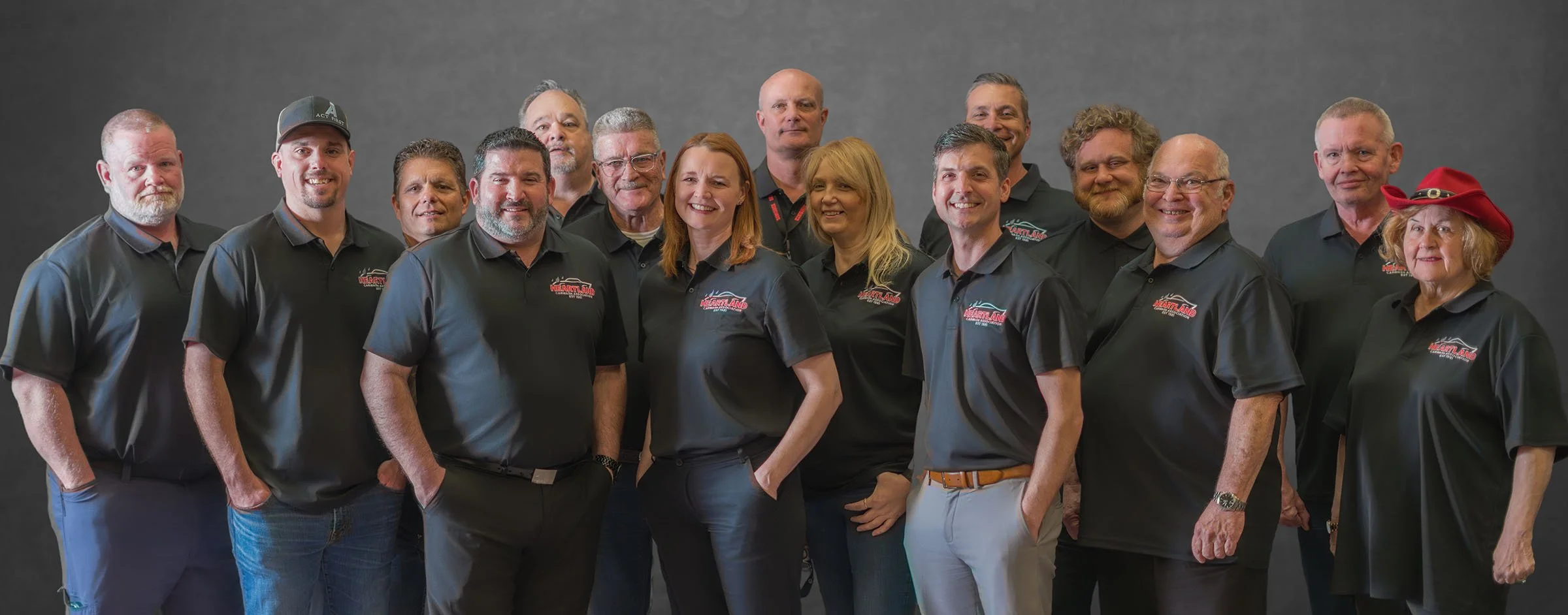 Group of 15 adults wearing black shirts with red and white logos, posing against a gray background, with one woman wearing a red hat.
