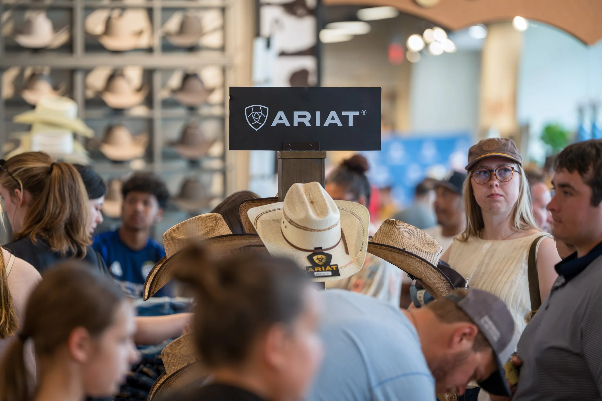 A display of various cowboy hats at an ARIAT store, with people shopping and looking at hats in the background.