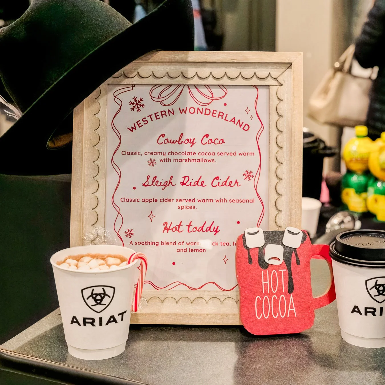 A menu board highlighting holiday beverages including Cowboy Coco, Sleigh Ride Cider, and Hot Taddy, with cups of hot chocolate and cider on the table, in a festive setting.