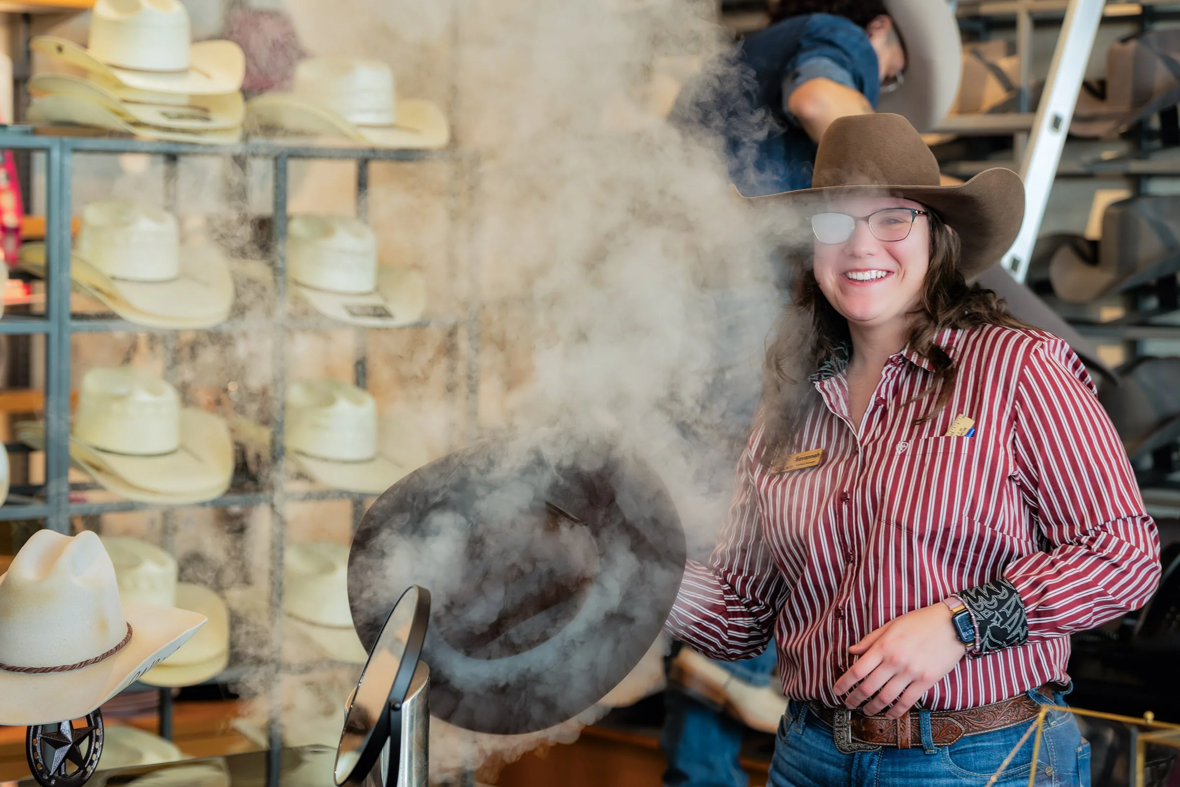 A woman wearing a cowboy hat, glasses, and a striped shirt is smiling while cooking with steam rising from a skillet in a hat store with shelves of hats around her.