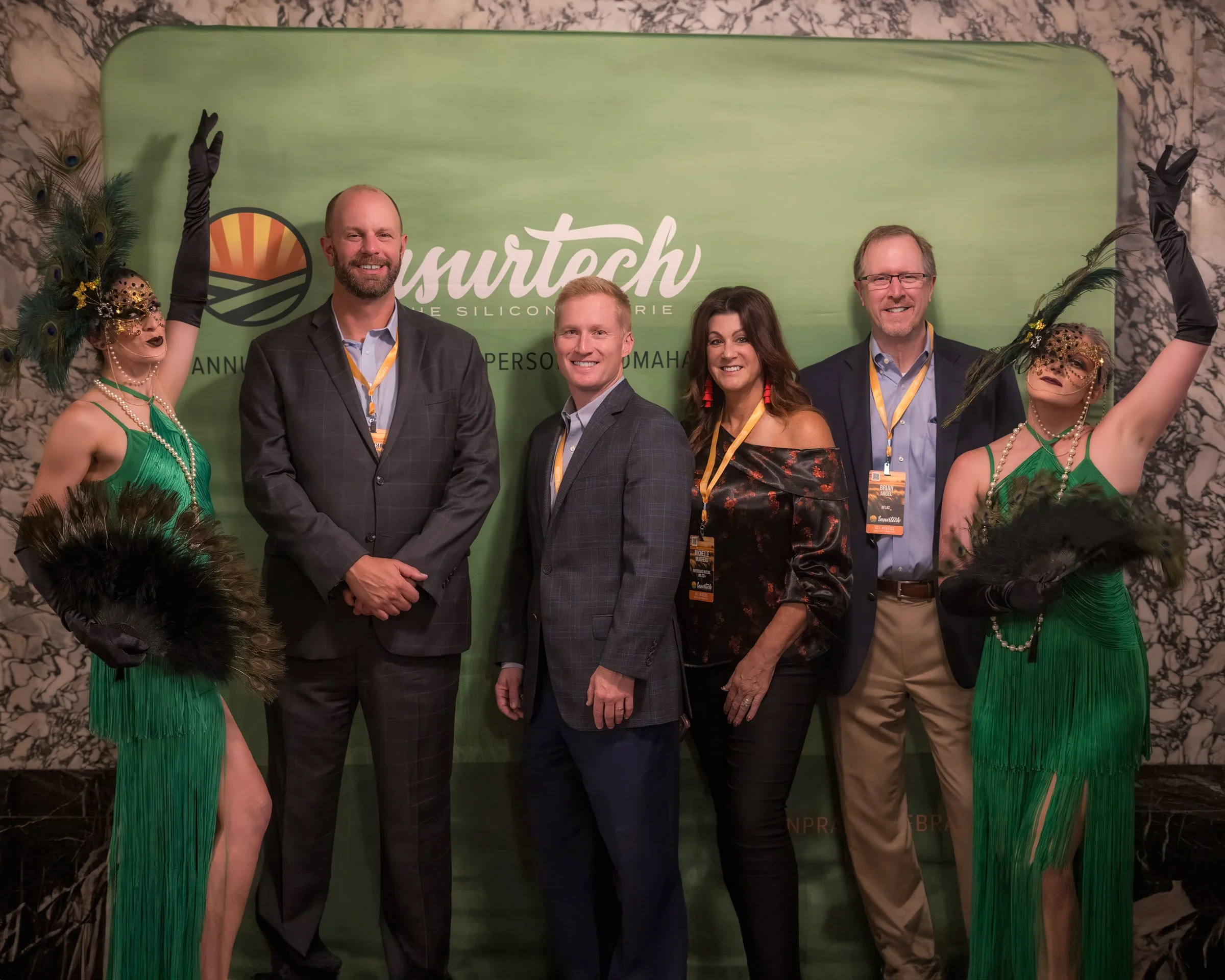 Group of five people posing in front of a green backdrop at an event, with two women dressed in green carnival costumes with peacock feather masks, and three men in business suits.