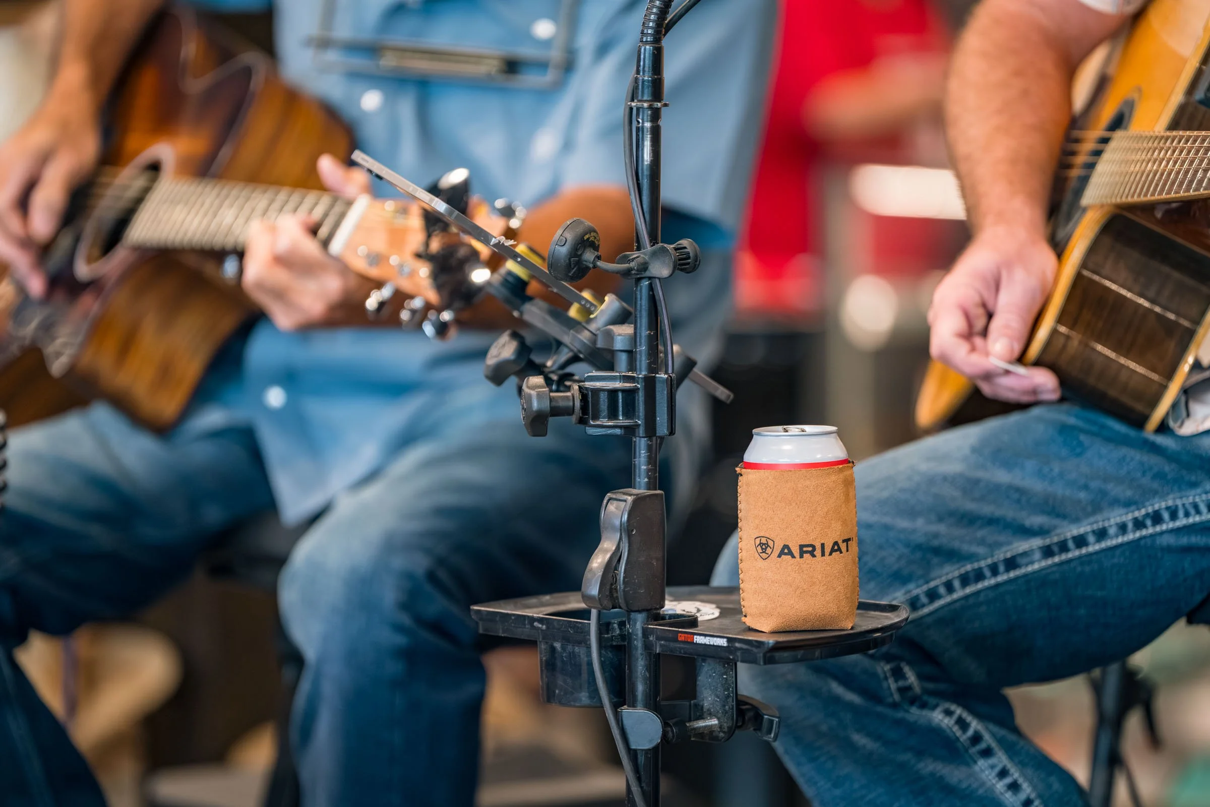 Two people playing acoustic guitars at an indoor event, with a microphone and a can of soda in a cozy setting.