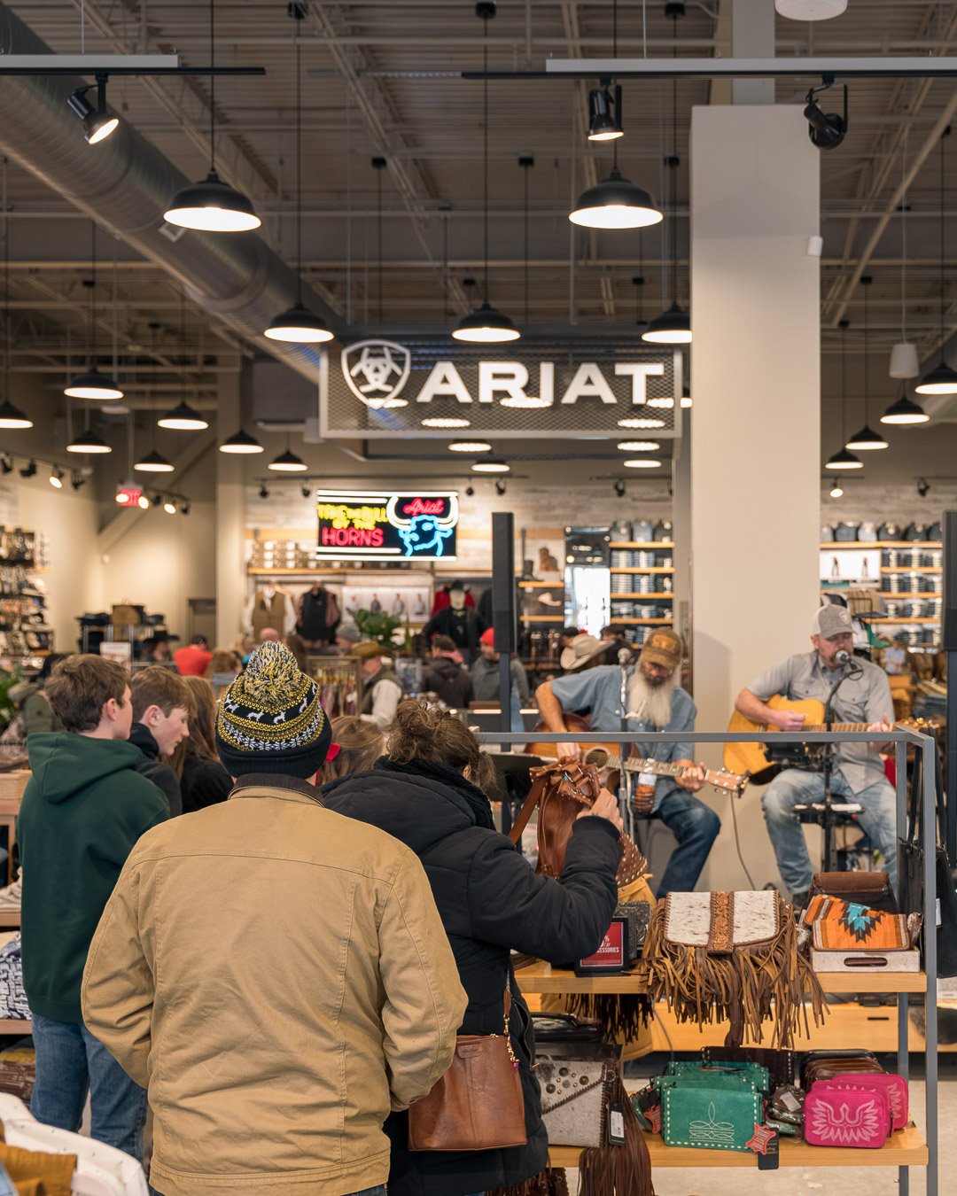 People shopping inside an outdoor gear store with a small live music performance happening in the background, featuring two musicians playing guitars.
