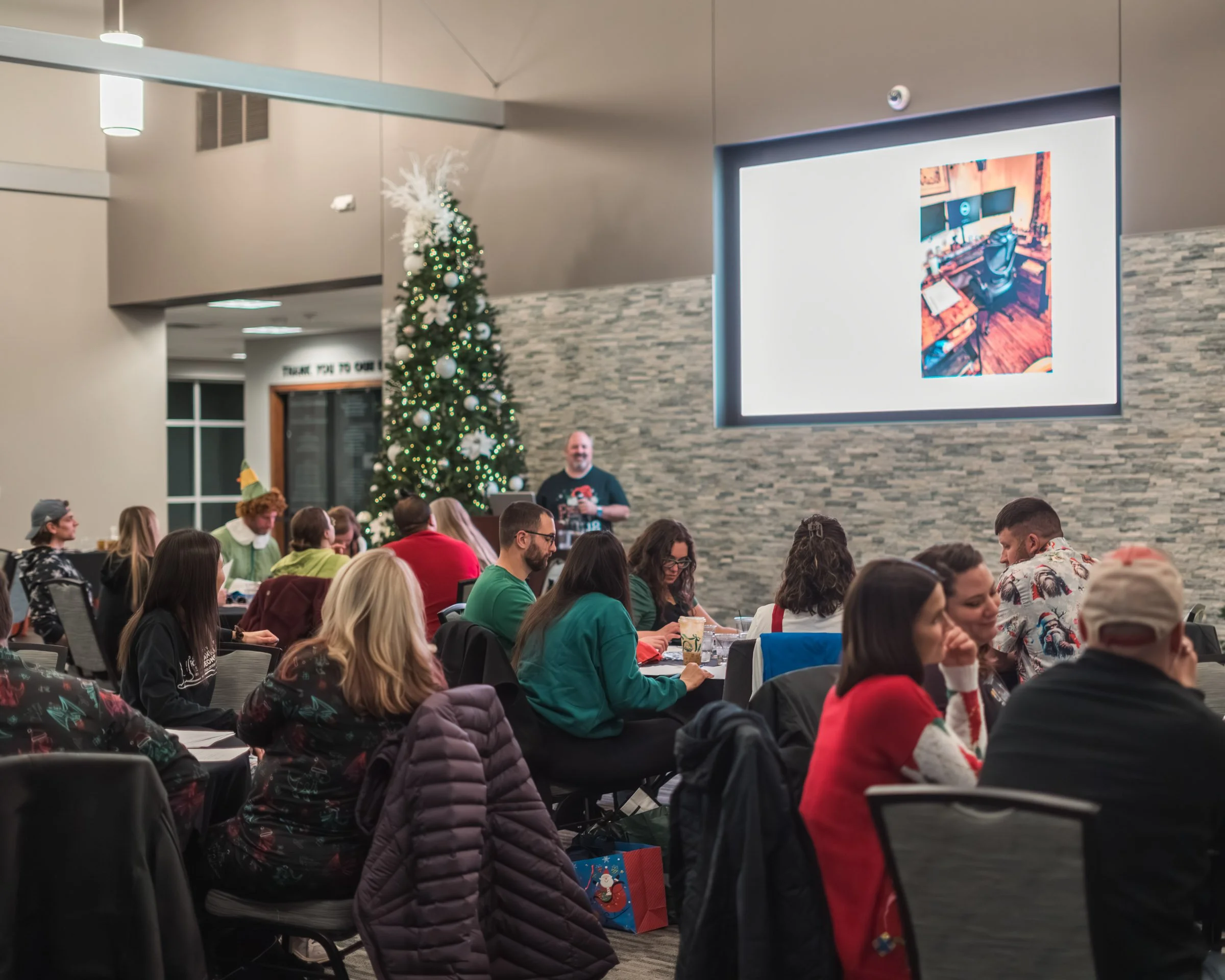 People gathered in a large indoor space for a festive event. There is a decorated Christmas tree with white ornaments and lights, and a man is speaking at a podium. A large screen displays an image of a workspace with dual monitors. The room has beig