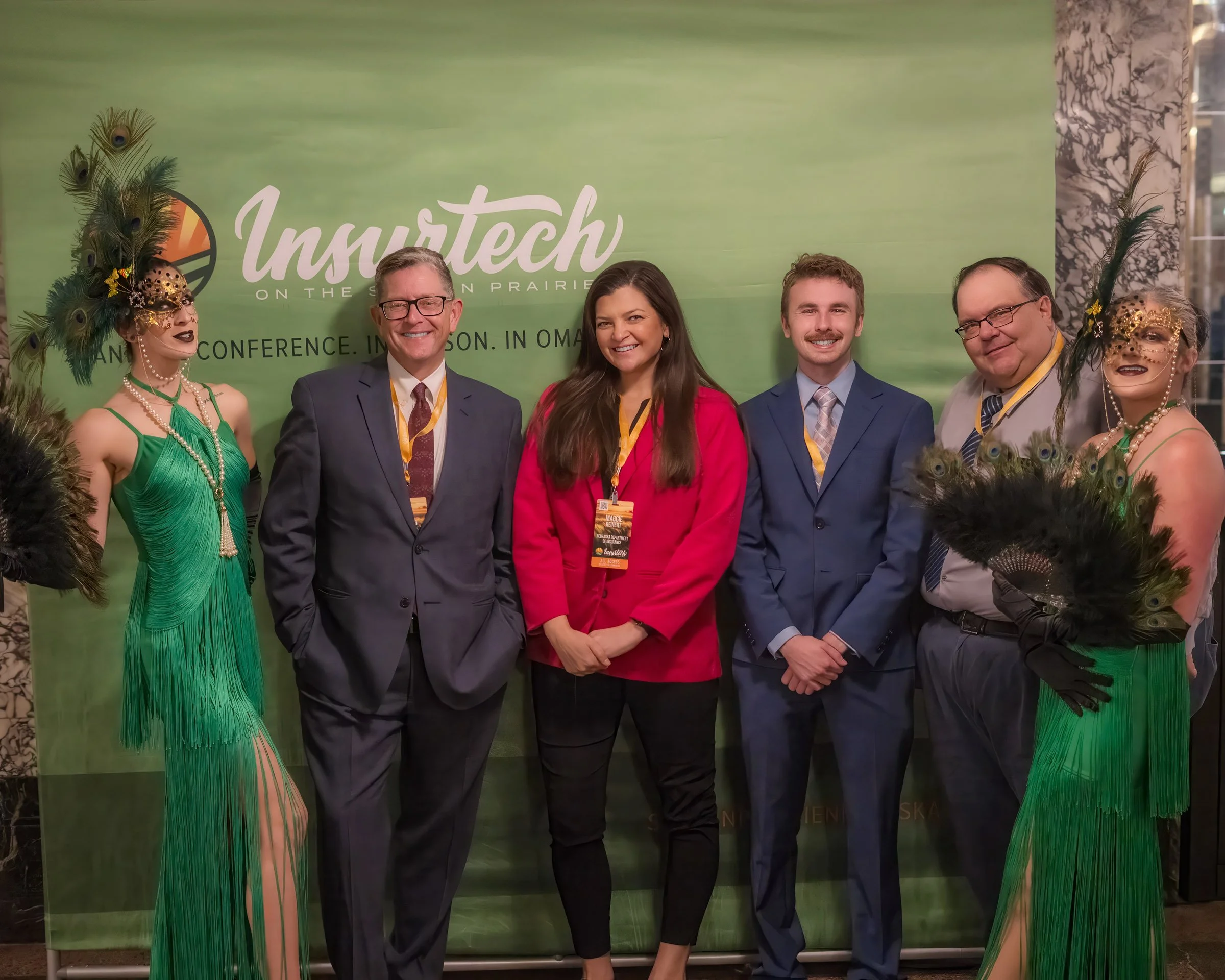 Group of people at a conference, including women dressed in green costumes with peacock feather accessories and people in business attire, standing in front of a green backdrop with the event logo 'Inustech'.