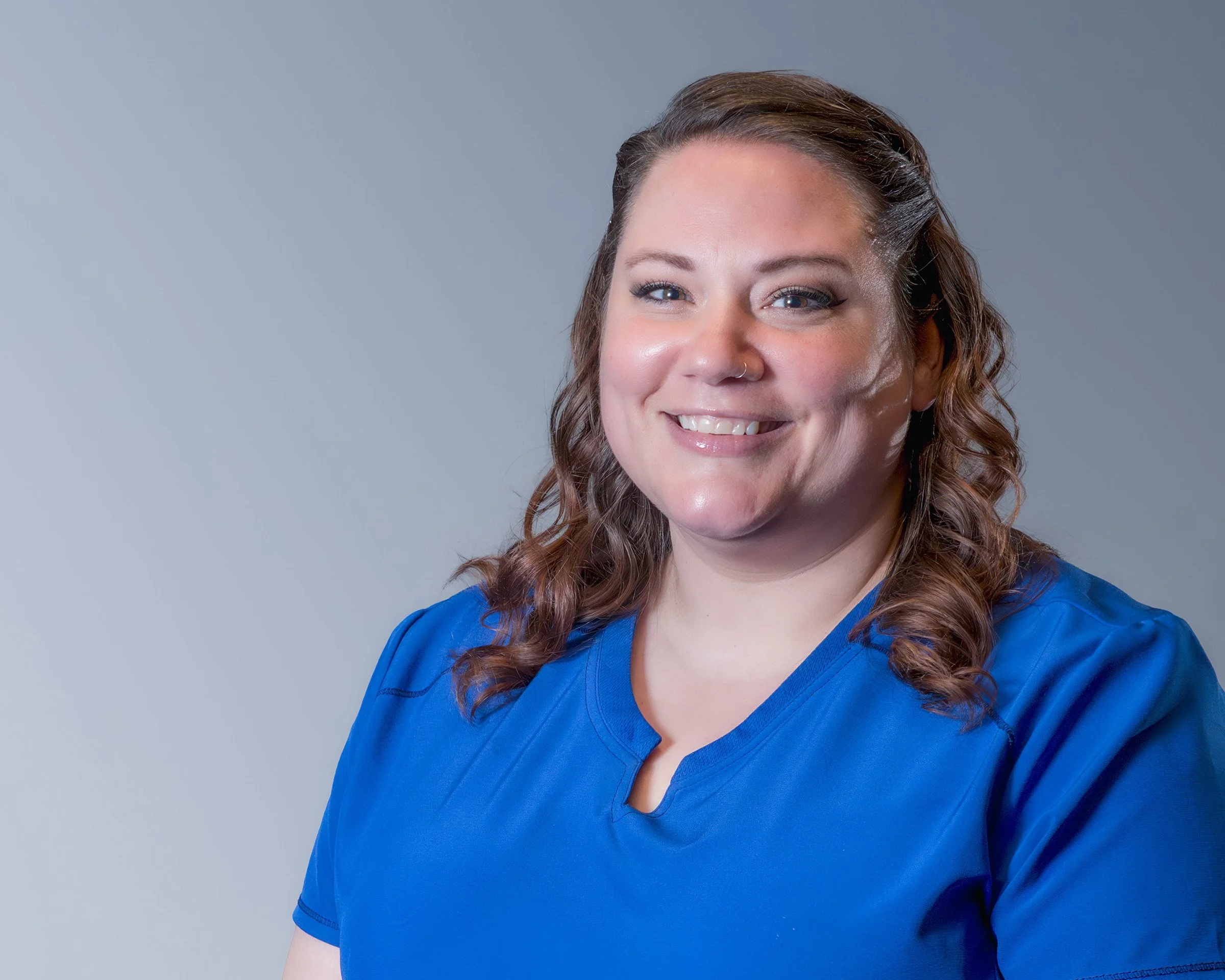 A woman with curly brown hair, blue eyes, and a nose ring, smiling while wearing a blue medical scrub top, against a gray background.