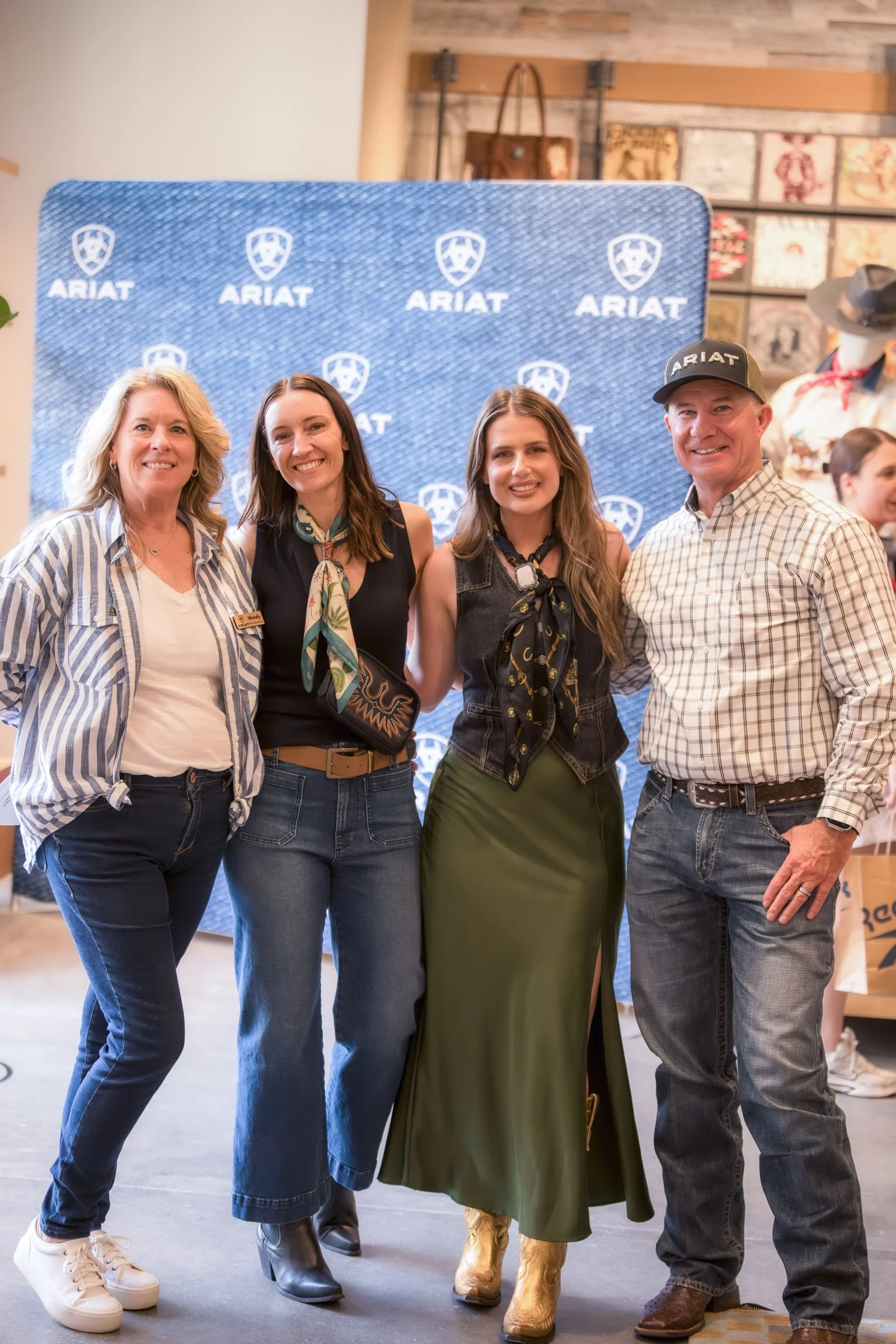 A group of four people posing in front of an ARIAT branded backdrop at an indoor event. They are smiling and dressed in casual Western-inspired attire, including boots and hats.