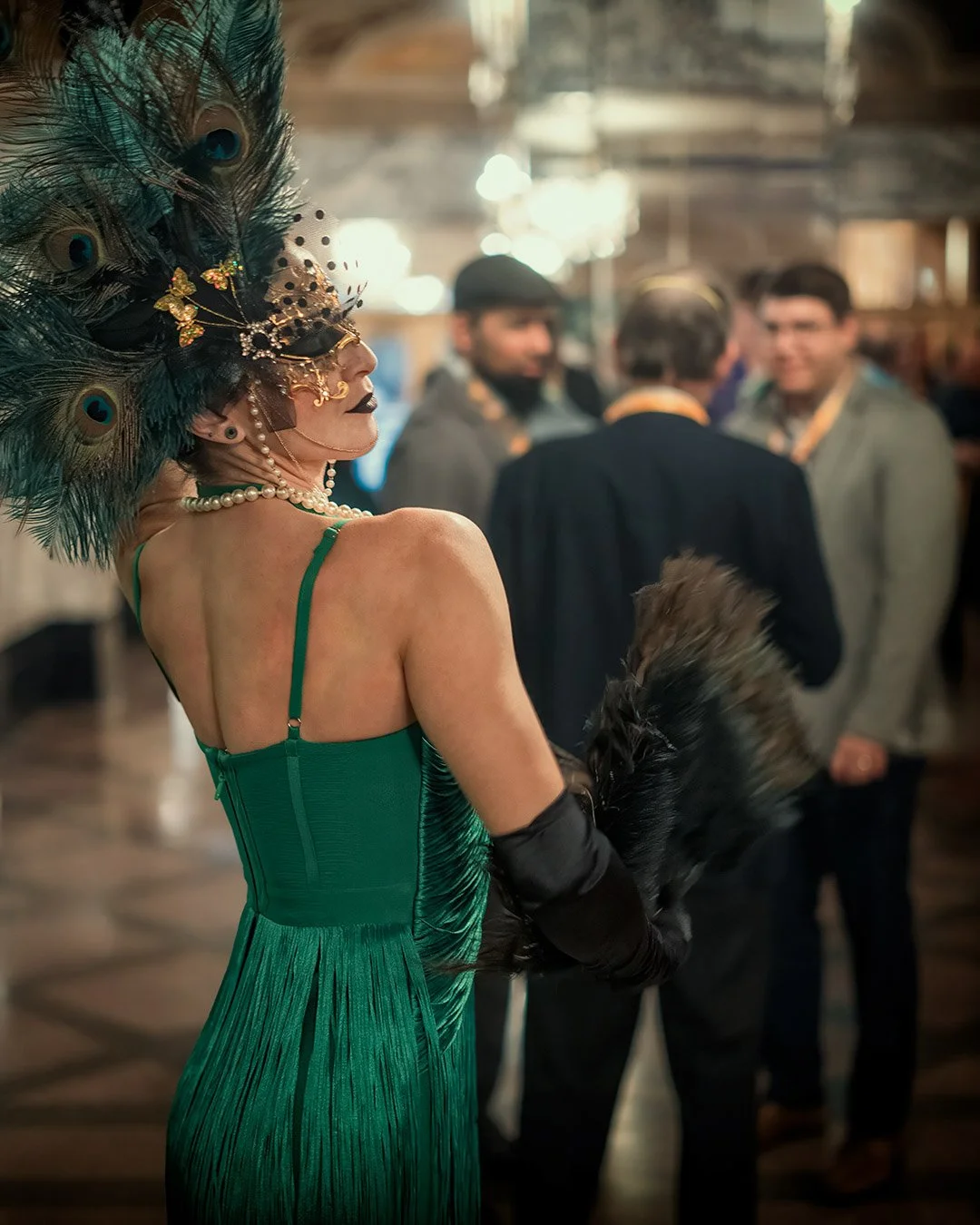 A woman in a green dress with a feathered headpiece and pearl necklace is at a social event, holding a black feathered fan, with a group of people talking in the background.
