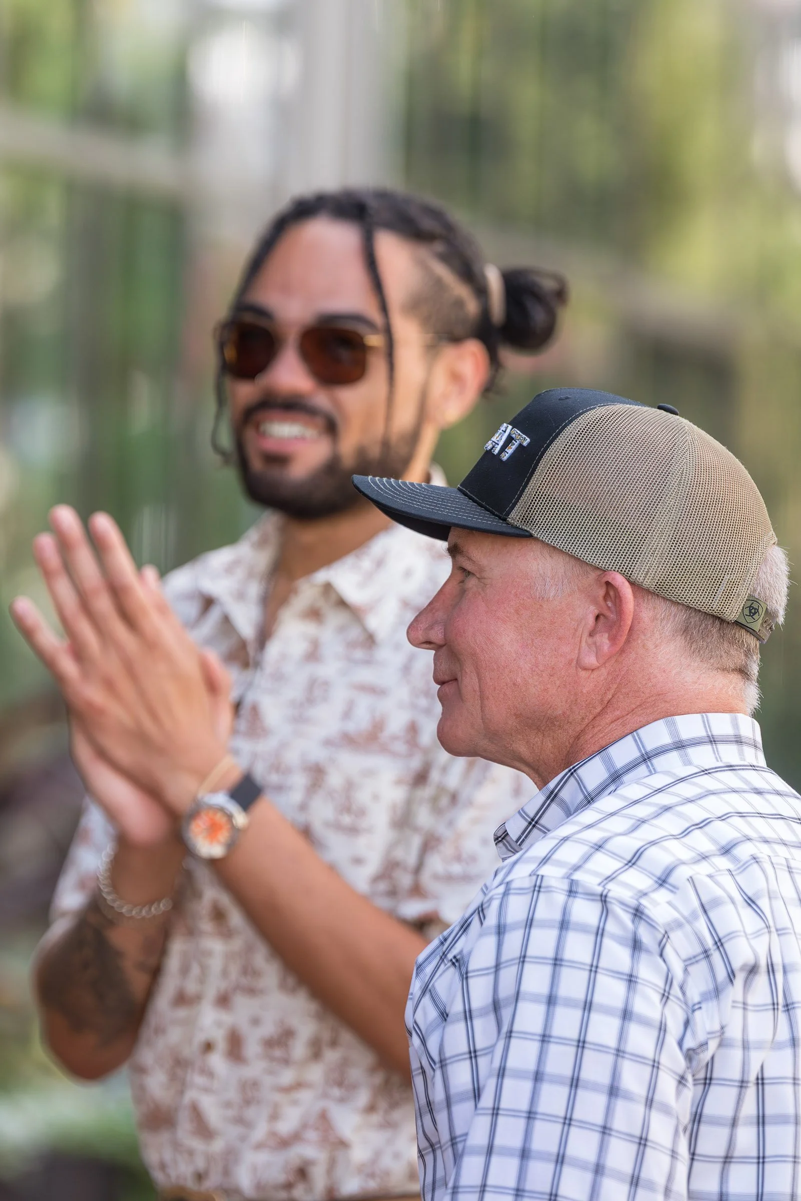 A man wearing a blue and white checkered shirt and a beige and black baseball cap is in the foreground looking to his left. Behind him, a man with long twisted hair, sunglasses, and a patterned shirt is smiling and clapping his hands. The background 