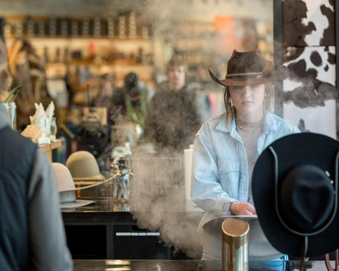 A woman wearing a cowboy hat and a striped shirt stands behind a counter, looking at a point of sale system, inside a cafe or bakery with shelves in the background. The photo is taken through a glass window with steam or smoke in the foreground.