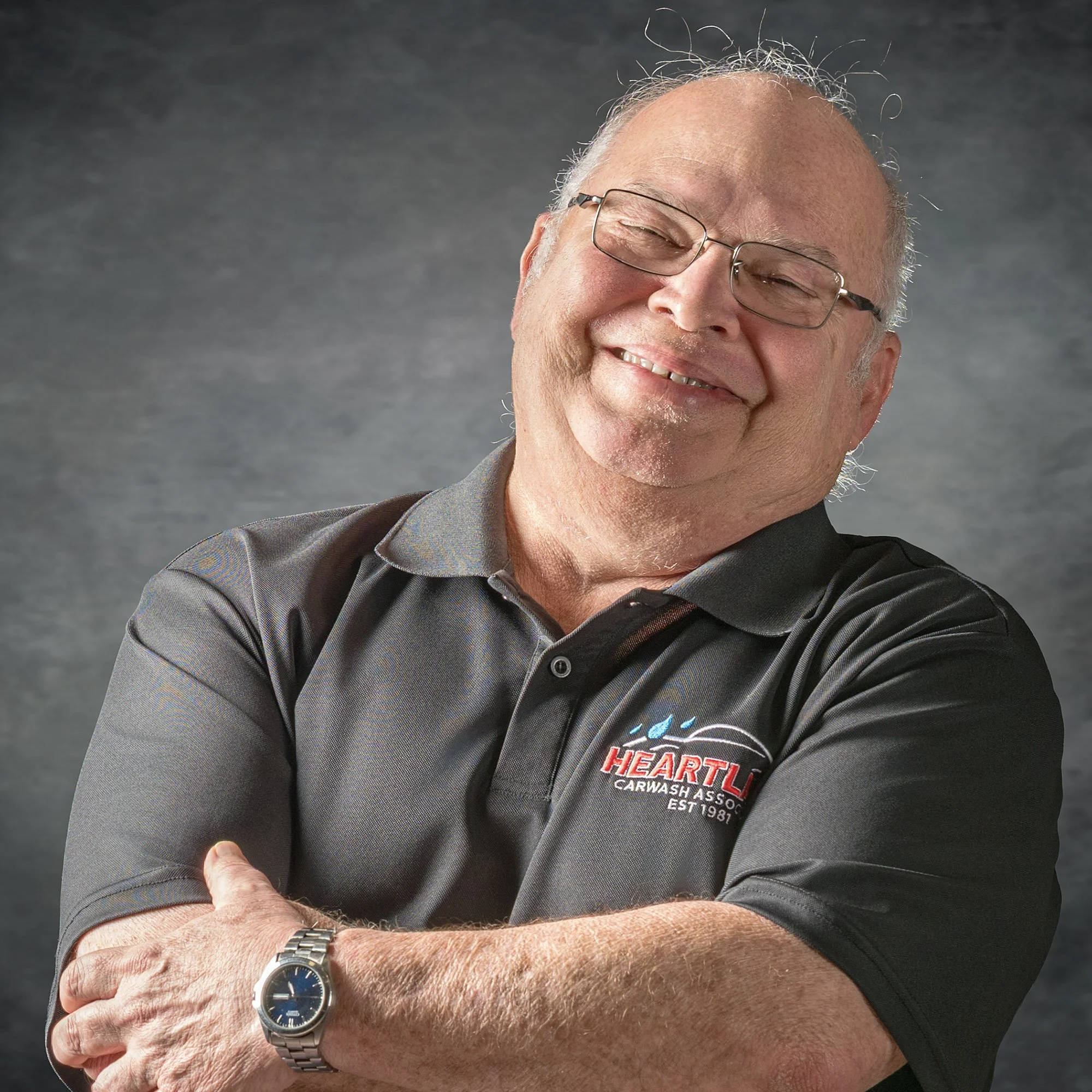 Close-up of an elderly man smiling, wearing glasses and a black baseball cap, showing a wristwatch on his left arm and an embroidered shirt with the logo of Hearts Auto Care.