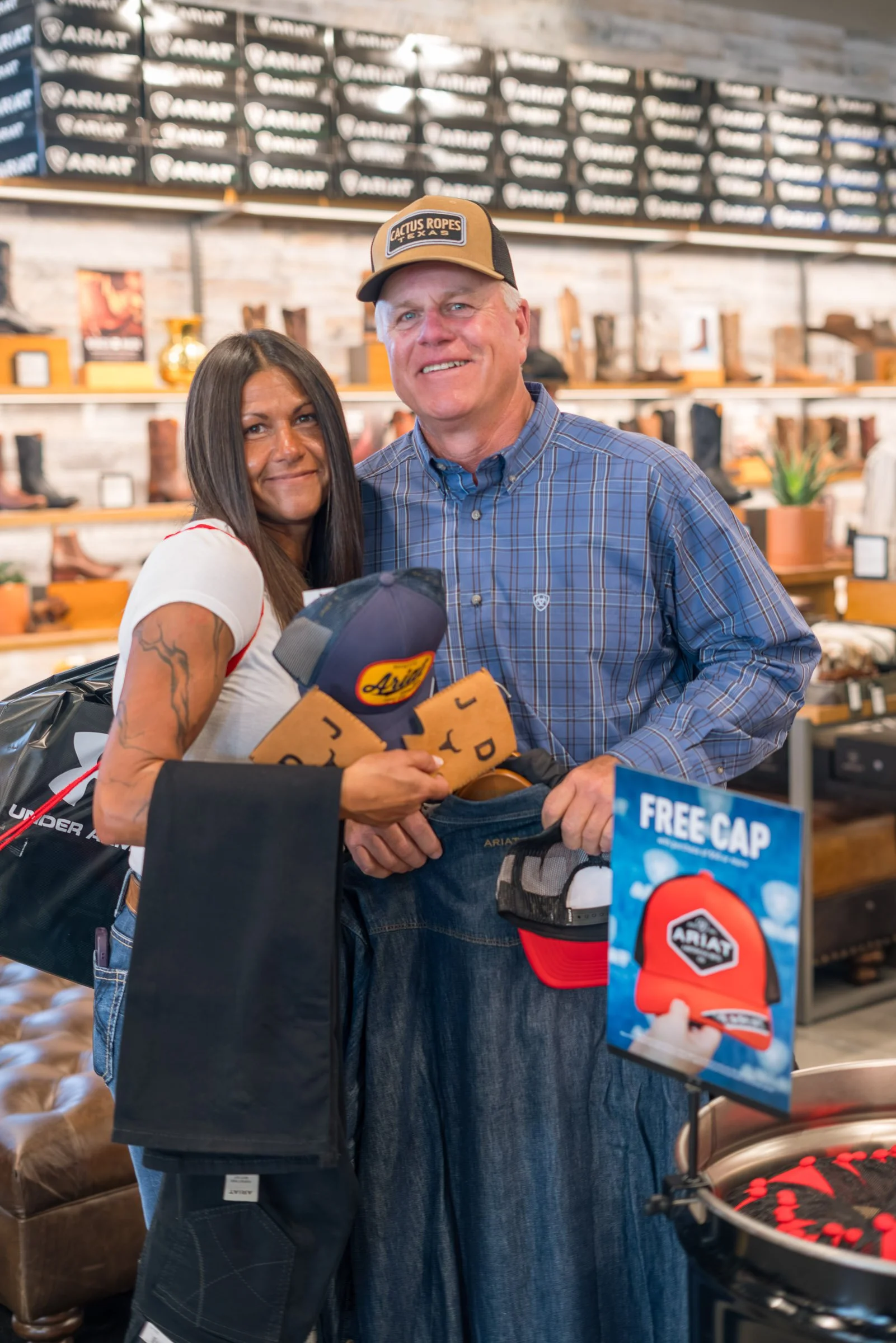 A man and woman shopping at a retail store, holding a hat and wearing casual clothing, with a display of boots and shoes in the background.