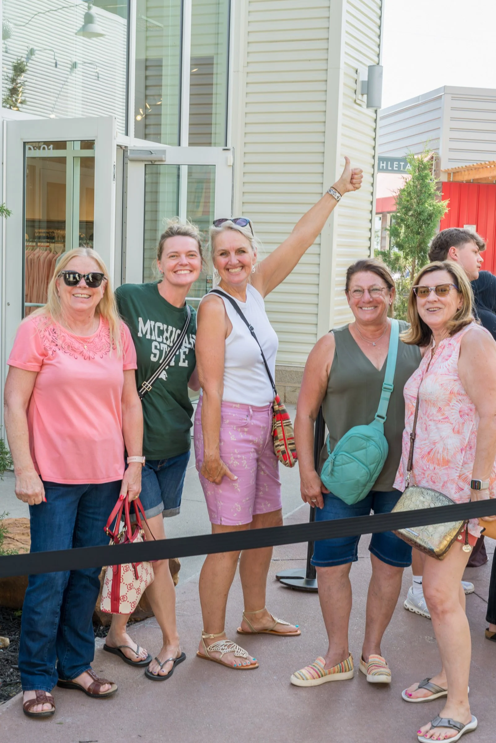A group of women standing outside in casual clothing, smiling and posing for the photo, with some women wearing sunglasses and sandals.