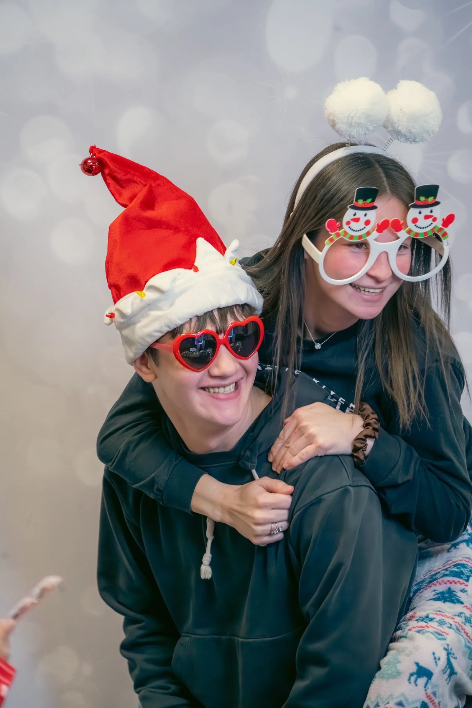 A woman and a child wearing holiday accessories, including a Santa hat and snowman glasses, smiling and hugging. The woman has white fluffy pom pom headbands, and the child has red heart-shaped sunglasses and a red Santa hat. Festive holiday spirit i