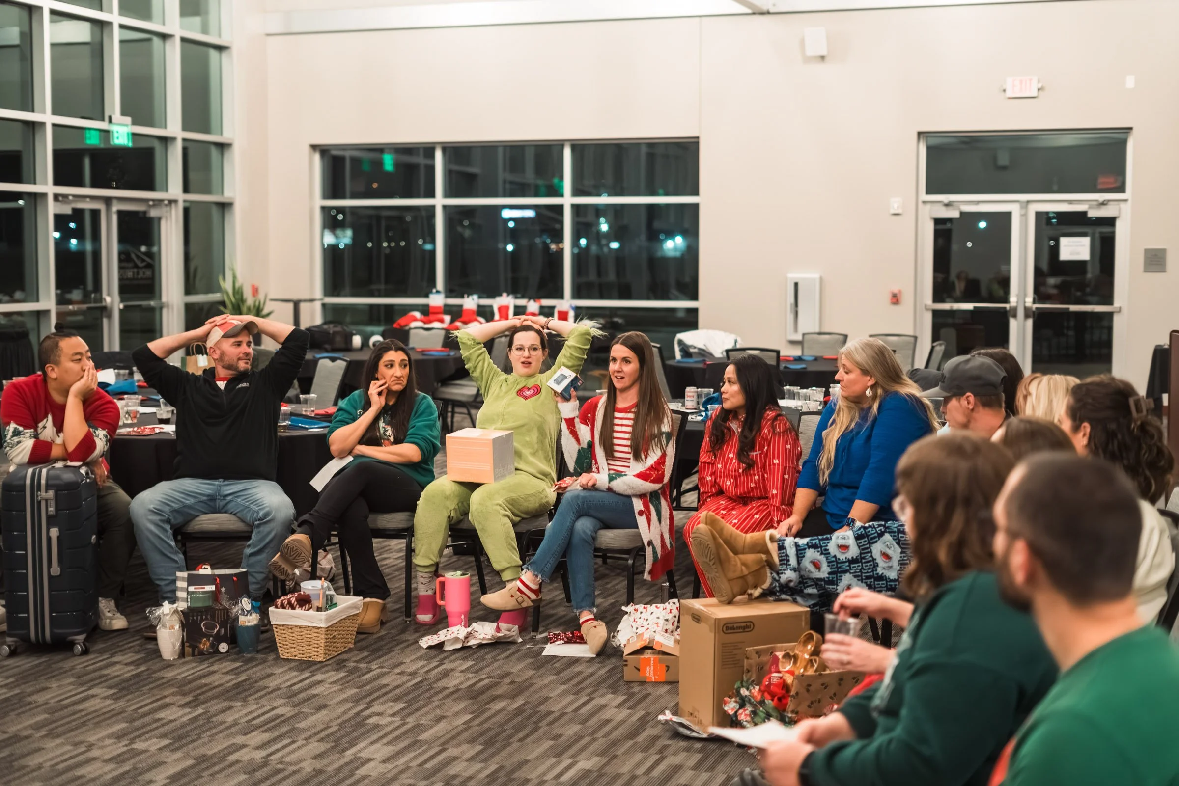Group of people sitting at tables decorated for Christmas, exchanging gifts, and wearing festive sweaters and pajamas. The setting is a large, modern indoor space with large windows and a high ceiling.