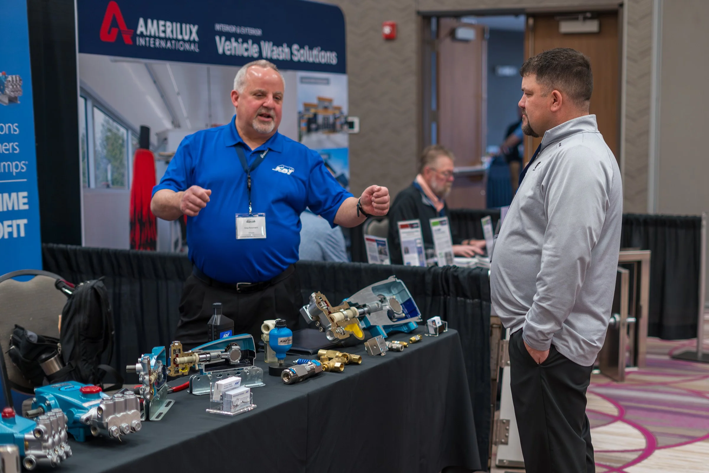 Two men are talking at a booth displaying various mechanical and plumbing parts at a trade show. One man is wearing a blue shirt with a name tag, and the other is dressed in a light gray jacket. There is a banner in the background that reads "Amerilux International Vehicle Wash Solutions."