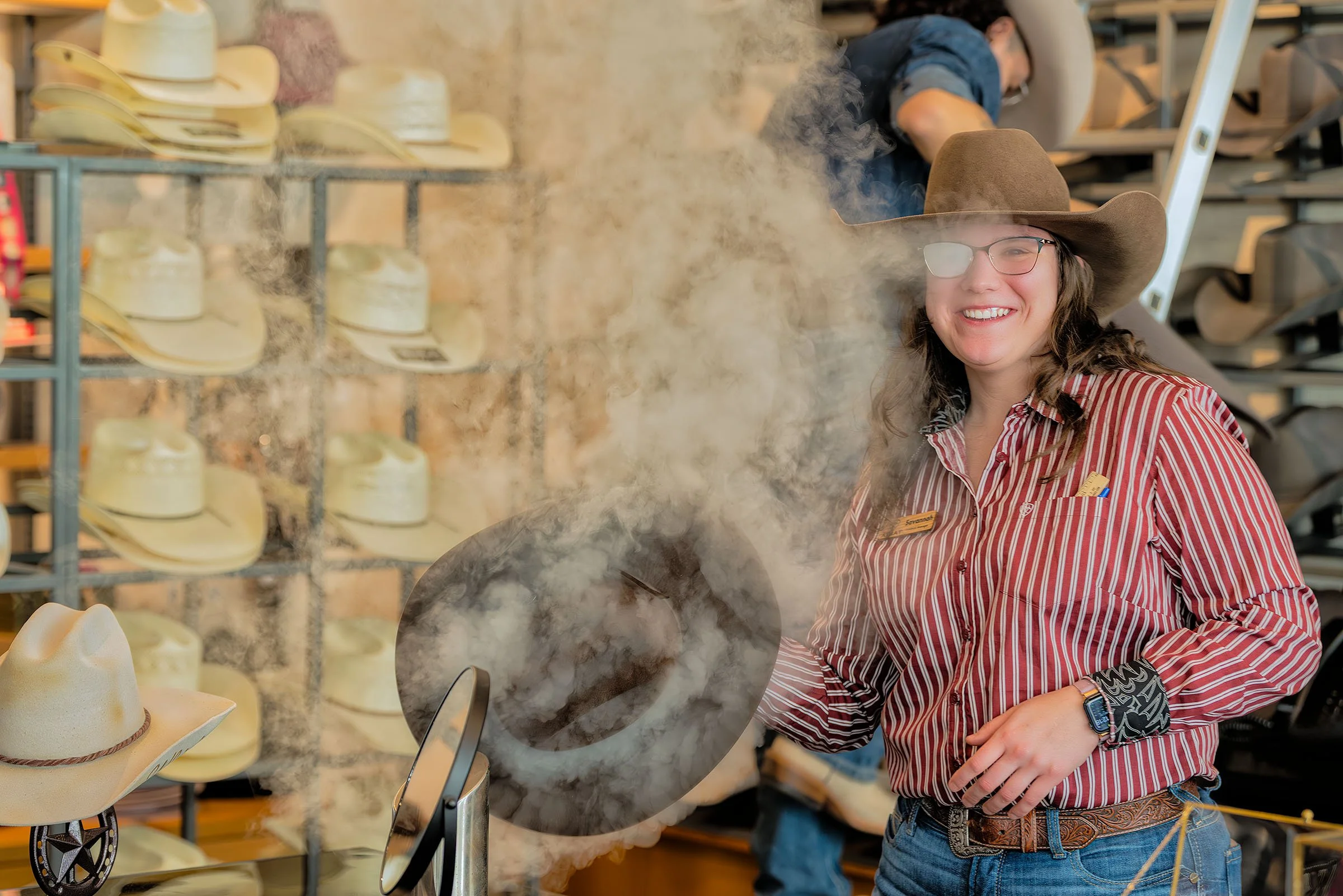 A woman wearing a cowboy hat, glasses, and a striped shirt, smiling while at a hat display in a store.