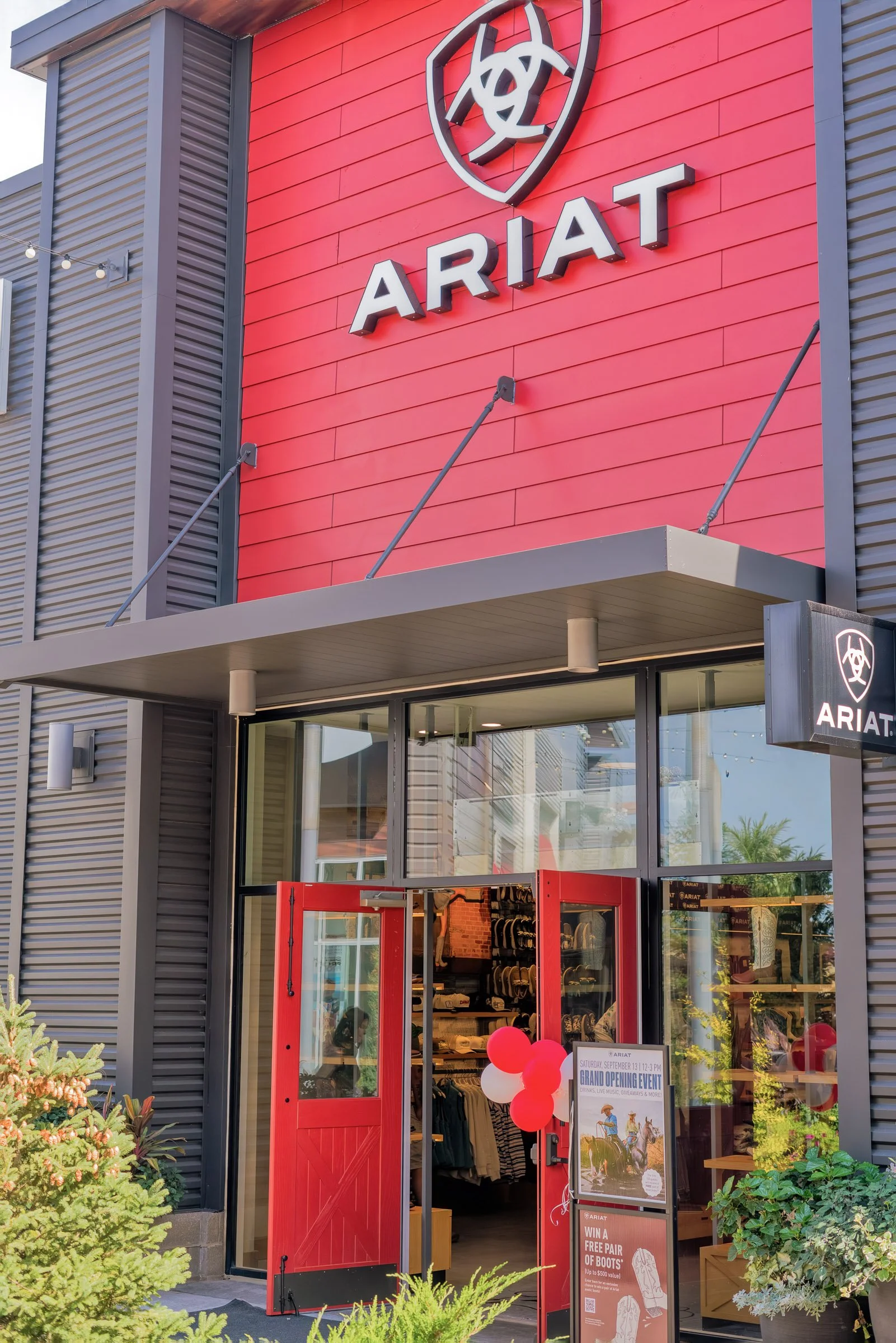 Exterior of a retail store called ARIAT, with a red door, large windows displaying hats and merchandise, and a sign announcing a grand opening event with balloons and promotional offers.