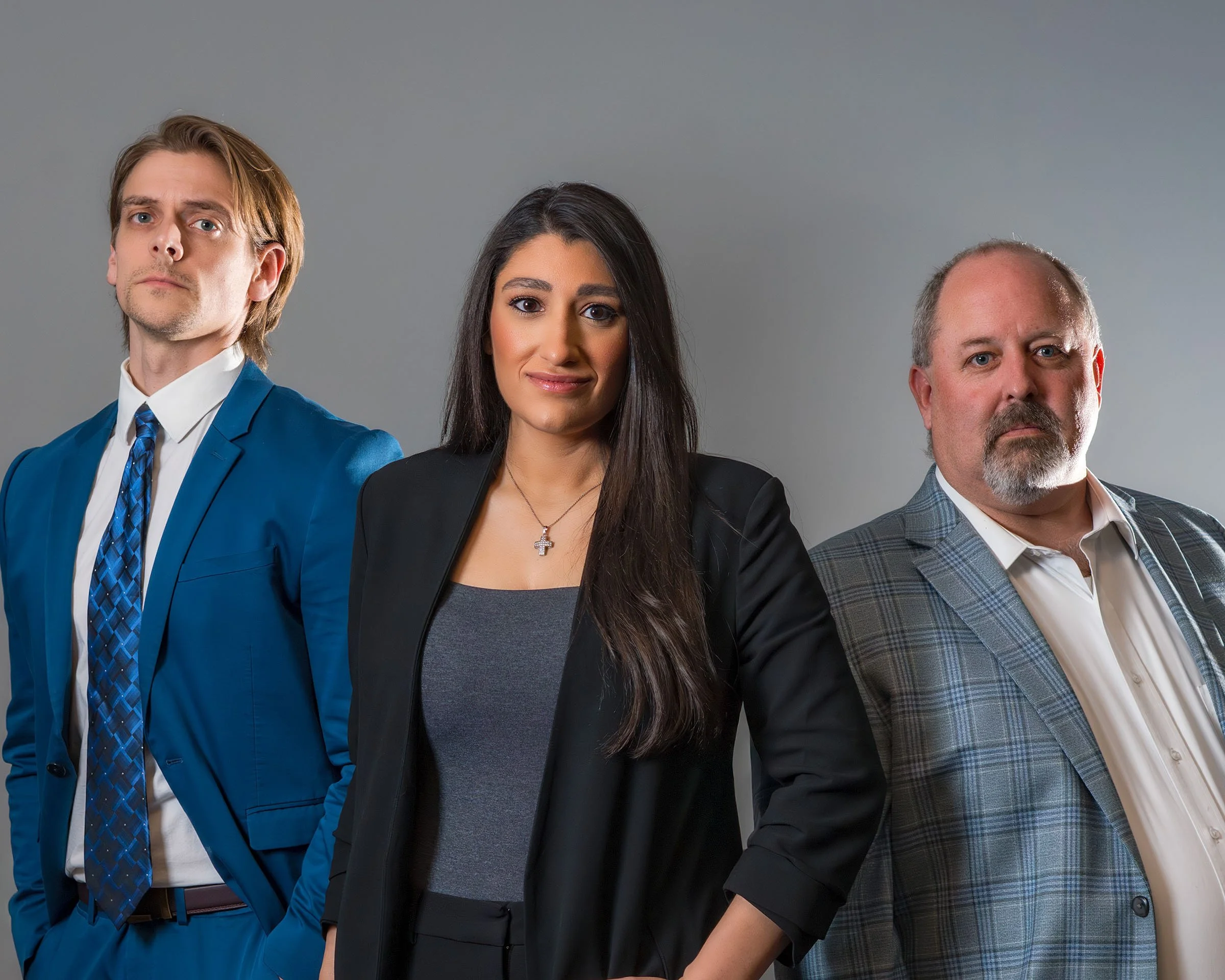 Three professionally dressed individuals standing against a neutral gray background, with the person in the middle slightly forward, the man to her left in a blue suit with a matching tie, and the man to her right in a checkered blazer and white shirt.