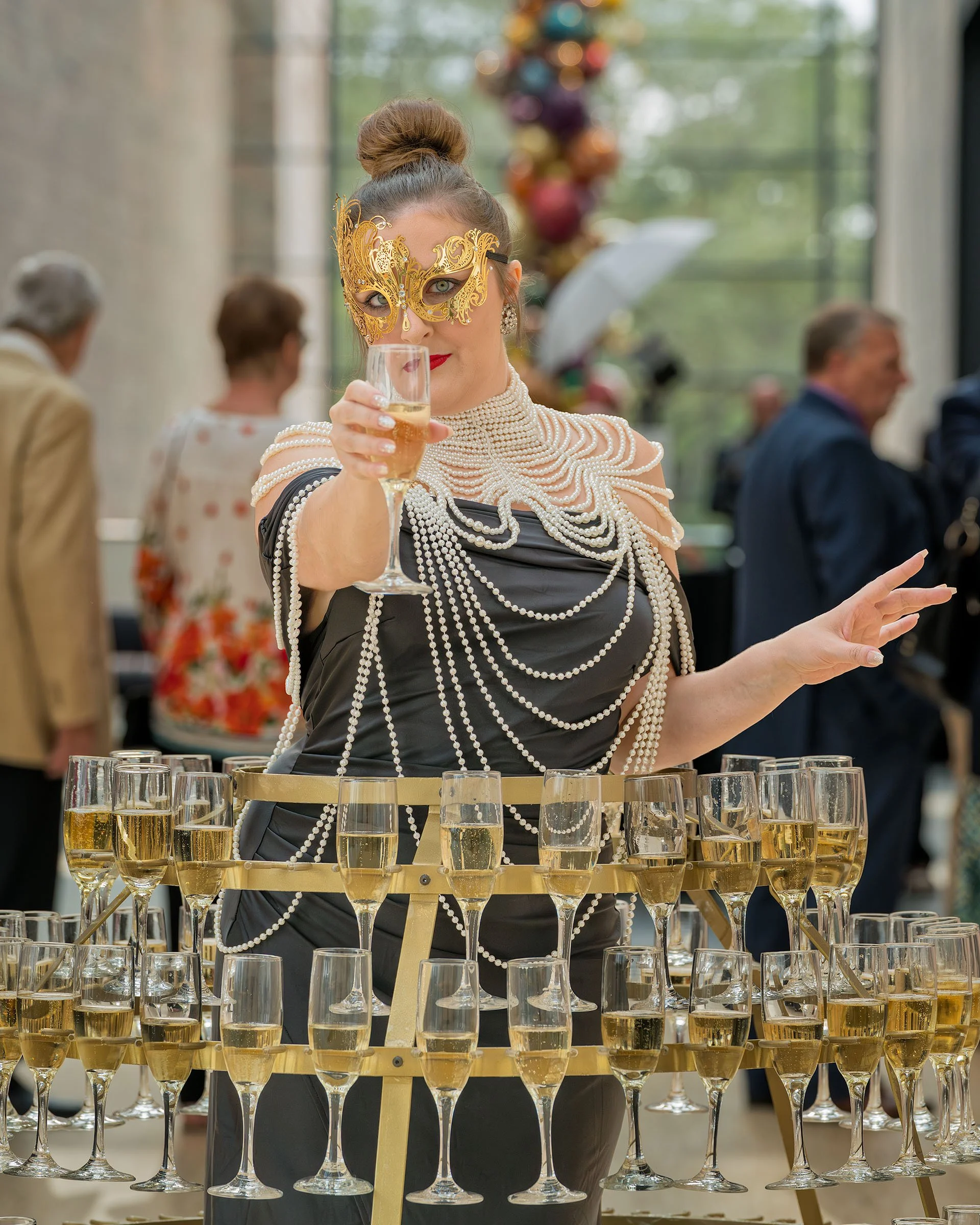 Woman dressed in elegant attire with pearl accessories and gold masquerade mask, holding a glass of champagne at a celebration with multiple glasses of champagne on a golden stand in front of her.