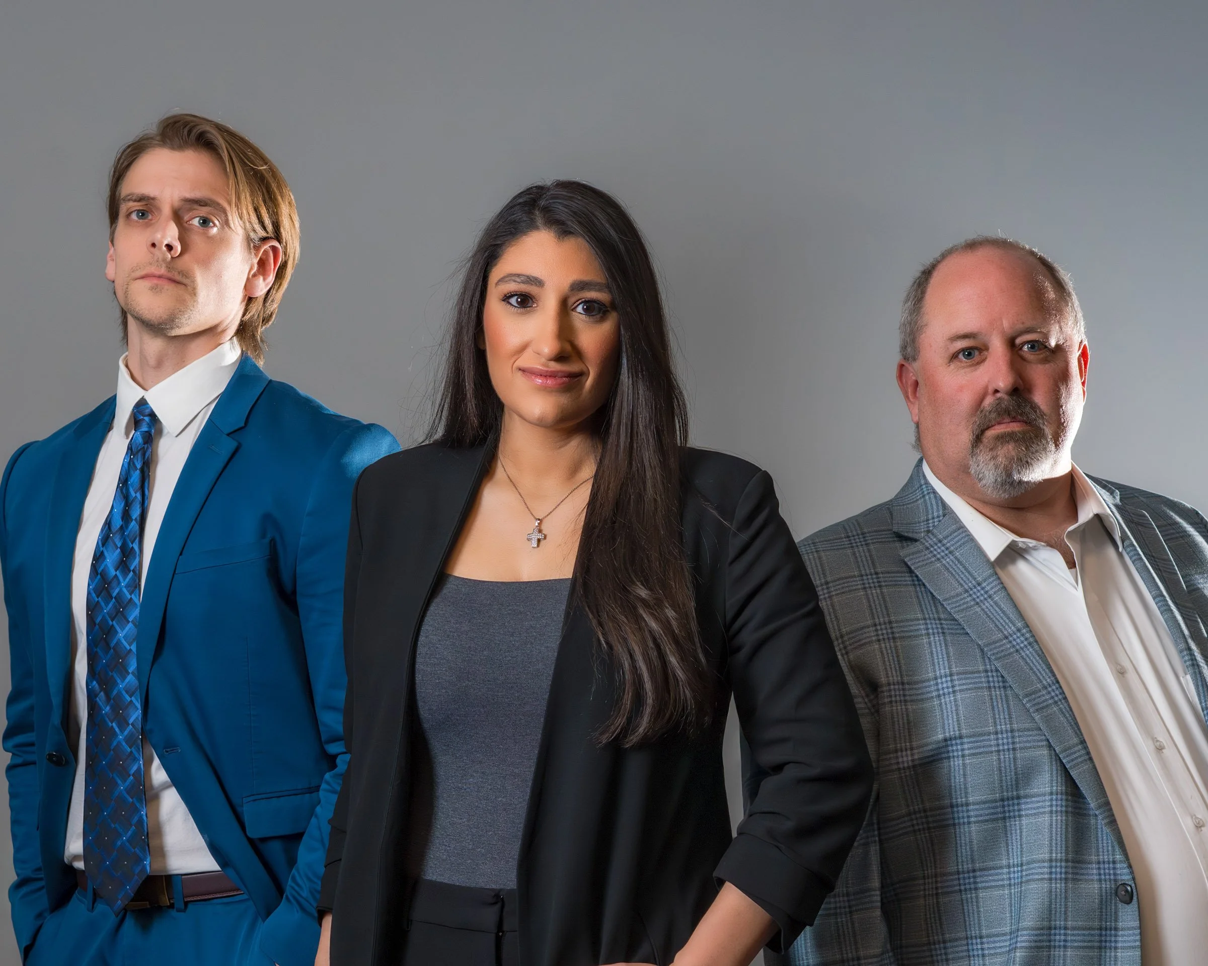 Three professionally dressed individuals standing side by side against a plain grey background.