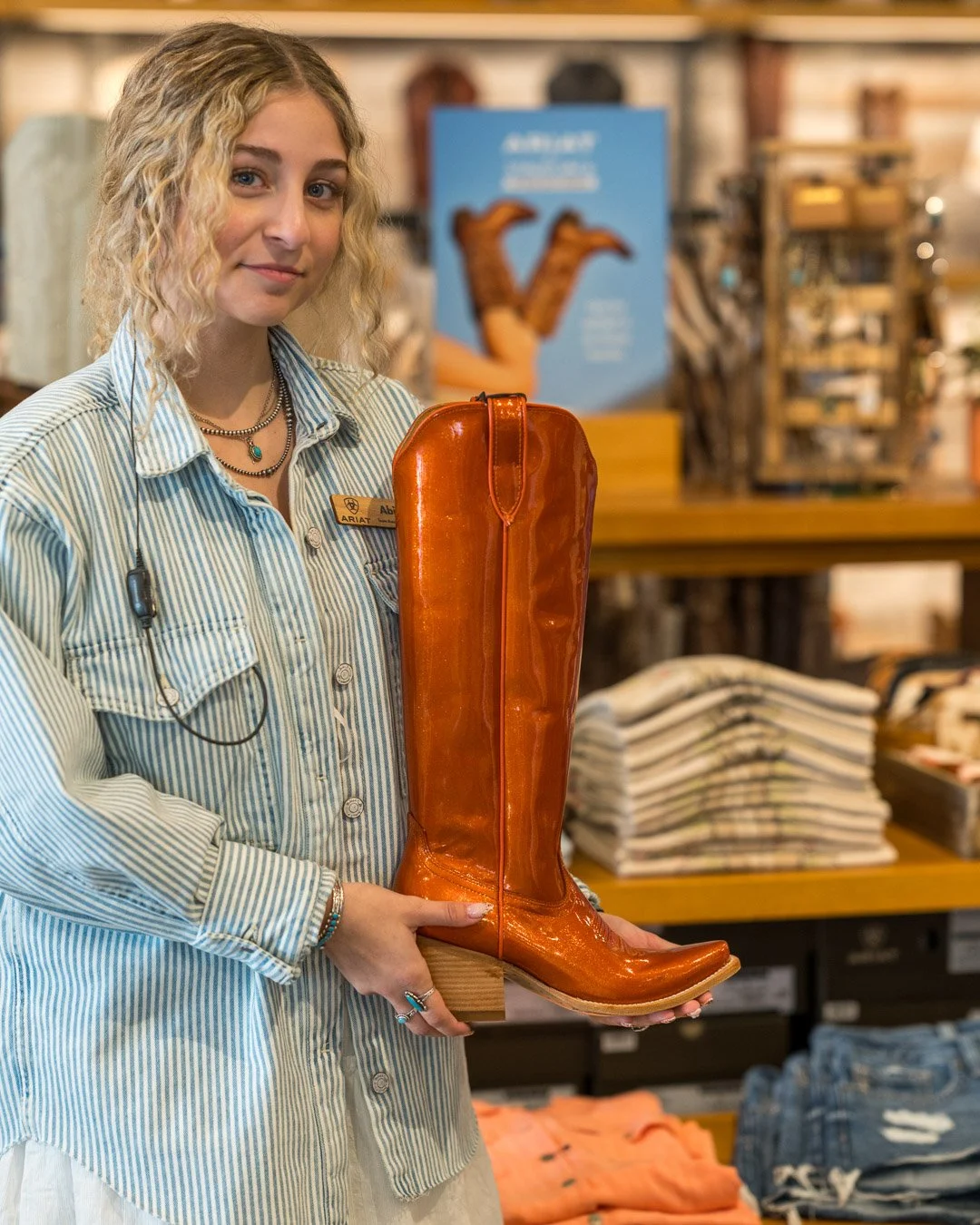 A woman with blonde curly hair wearing a striped shirt and multiple necklaces is holding a shiny orange cowboy boot display. She has a name badge that reads 'Abby'. The background shows stacked folded clothes and a blue poster with a cowgirl image.
