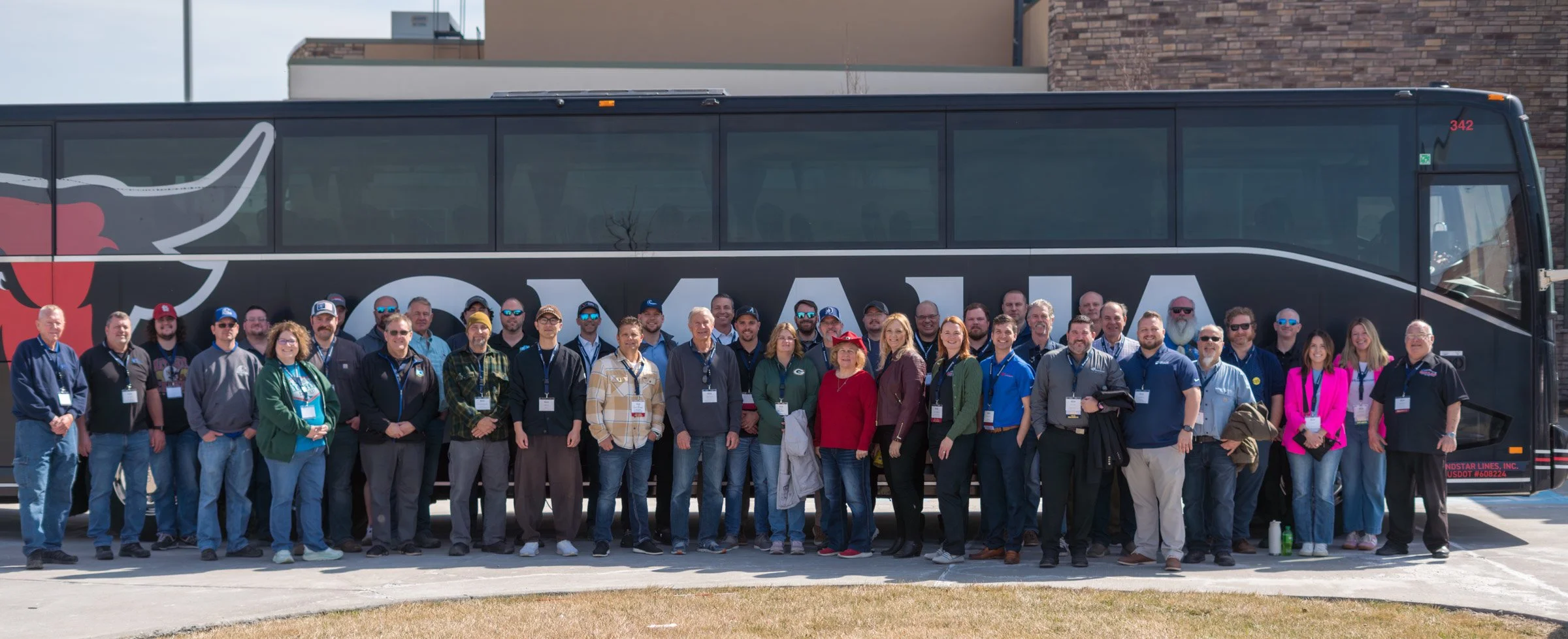 Group of diverse people standing in front of a large black bus with a red and white logo, outdoors on a sunny day.