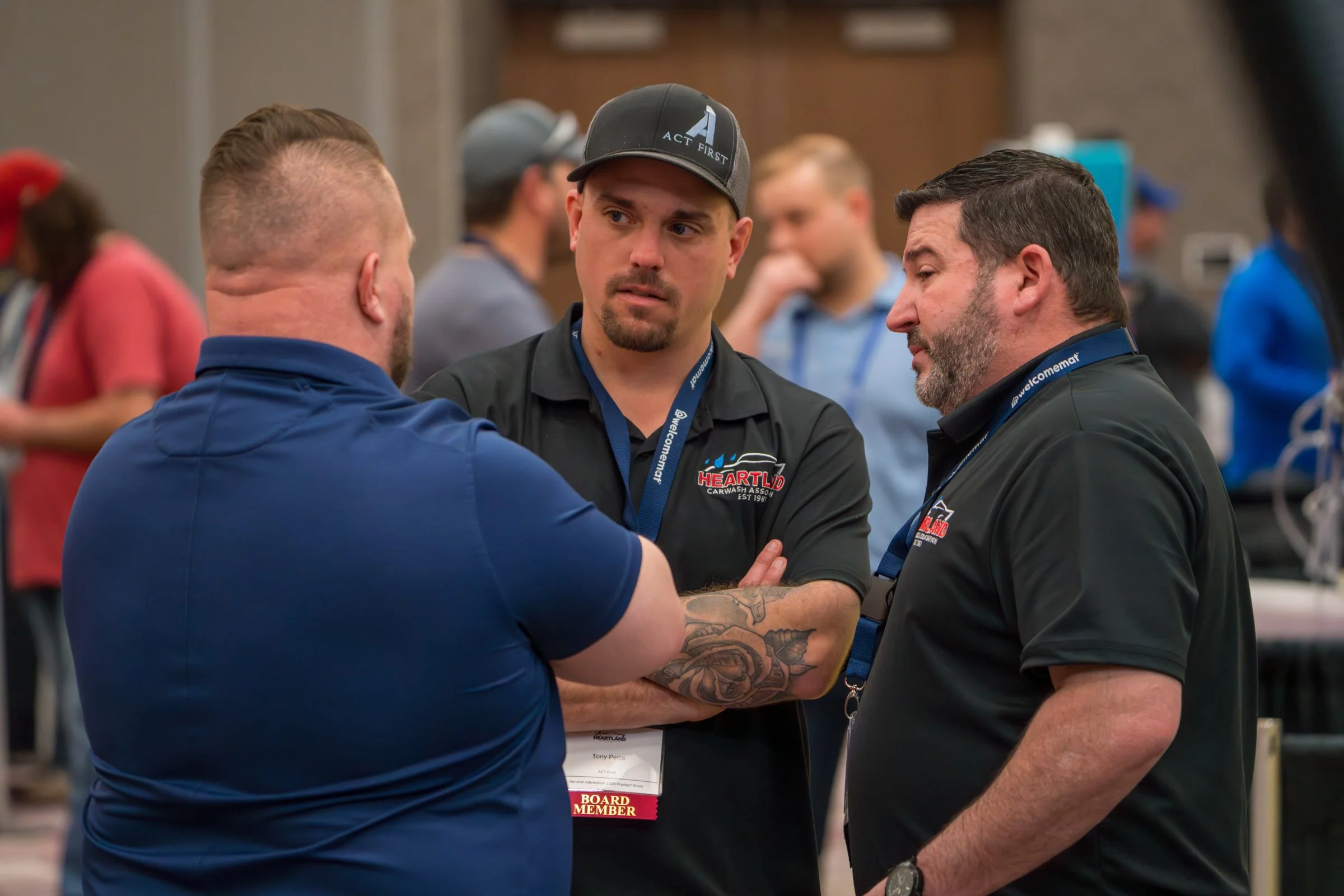Three men are engaged in a conversation at a conference or event, with one man wearing a shirt that says 'Heartland Carwash Association' and a badge that indicates he is a board member. They are standing closely, and others are visible in the backgro