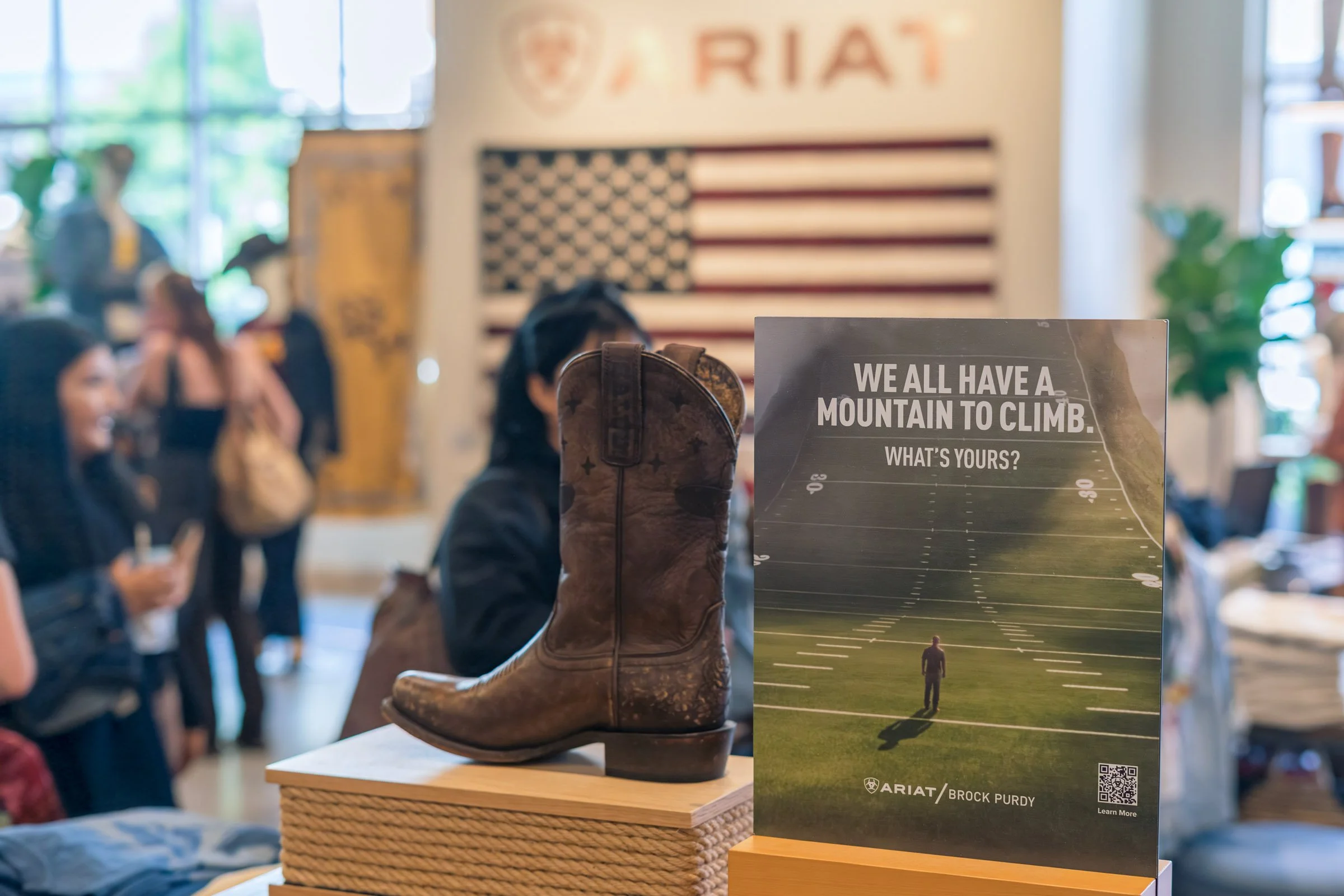 A vintage brown leather cowboy boot with a worn look is displayed on a small wooden table. Behind it, there is a poster with the text "We all have a mountain to climb. What's yours?" and an image of a person climbing a hill or mountain. In the backgr