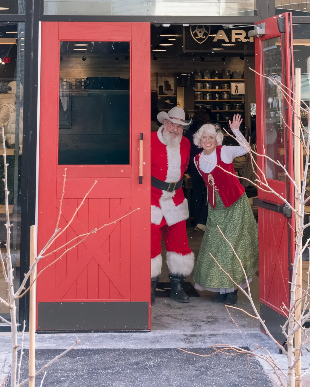 People dressed as Santa Claus and Mrs. Claus standing in a store doorway, smiling and waving during the holiday season.